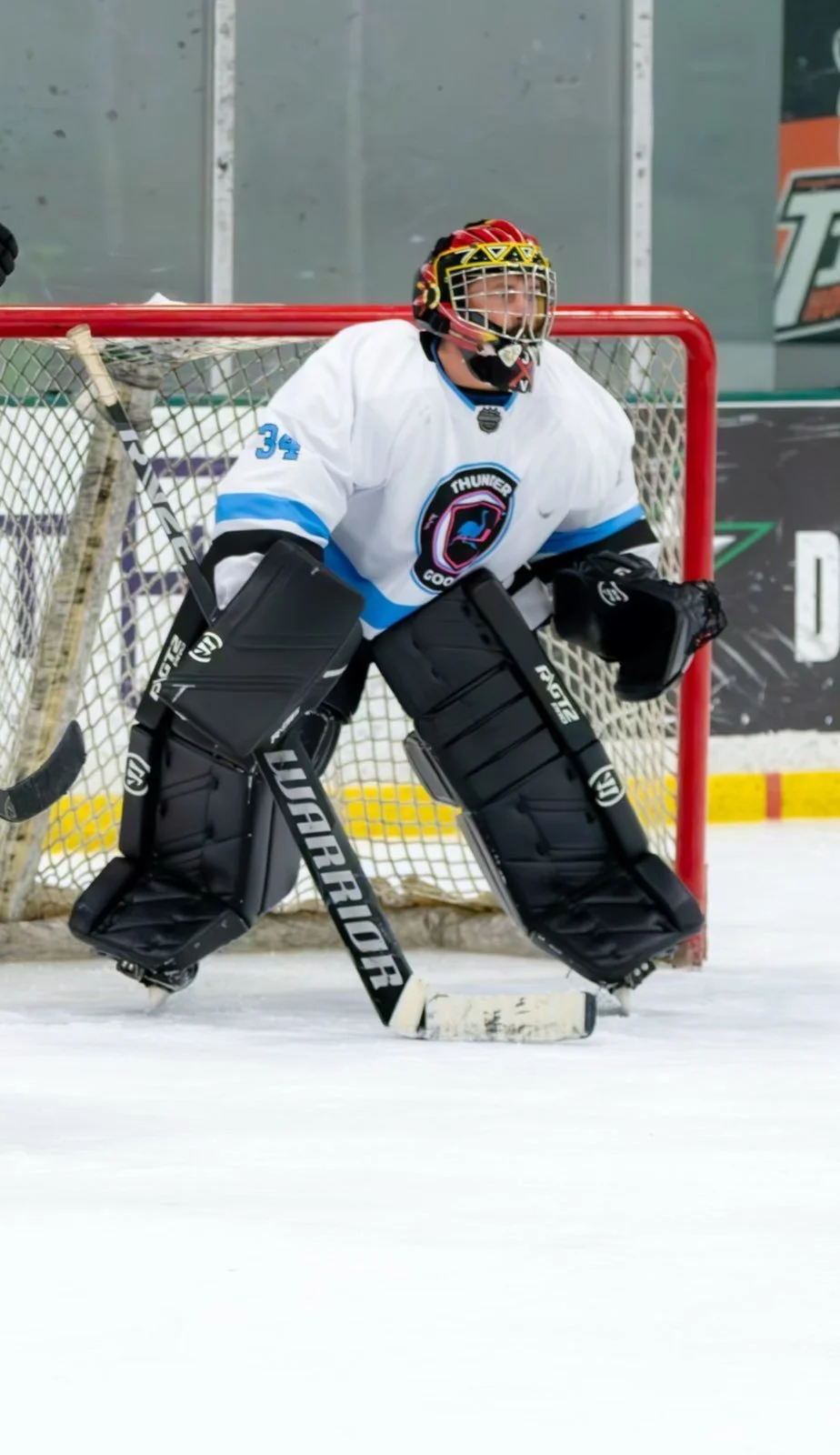 Ice hockey goalie in white jersey with blue accents, standing in front of the net, wearing protective gear including leg pads, blocker, and mask.