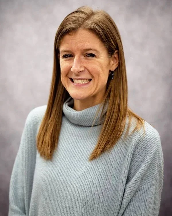A woman with light brown hair wearing a light gray turtleneck sweater, smiling against a plain gray background.