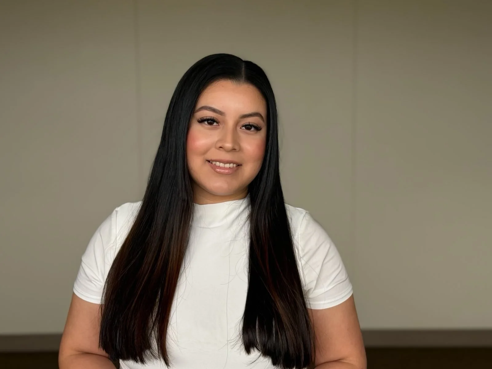 A woman with long black hair, wearing a white top, smiling at the camera against a plain wall background.