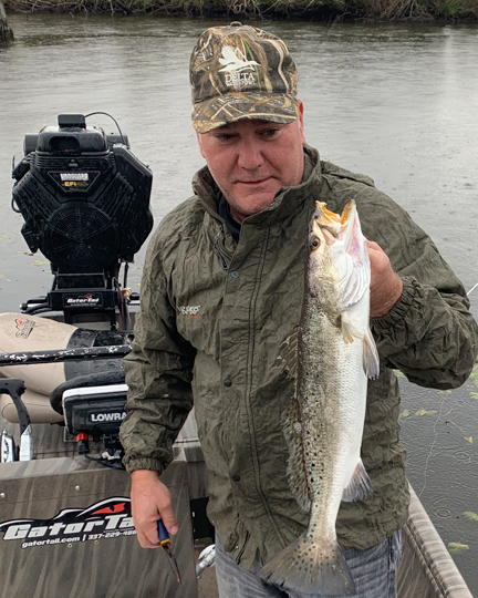 A man in outdoor clothing holding a large fish, standing on a boat in a river.