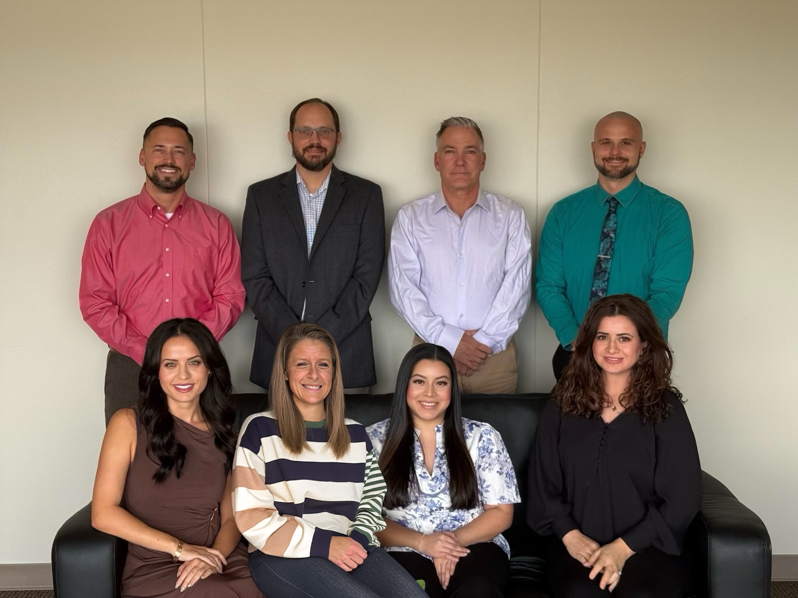 A group of nine people, four men standing in the back and five women sitting in front, posing for a photo in an office setting.