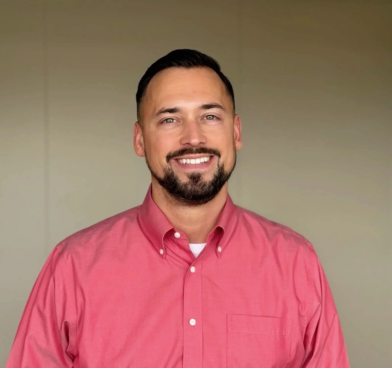 A man with dark hair and a beard smiling, wearing a pink dress shirt, standing against a neutral background.