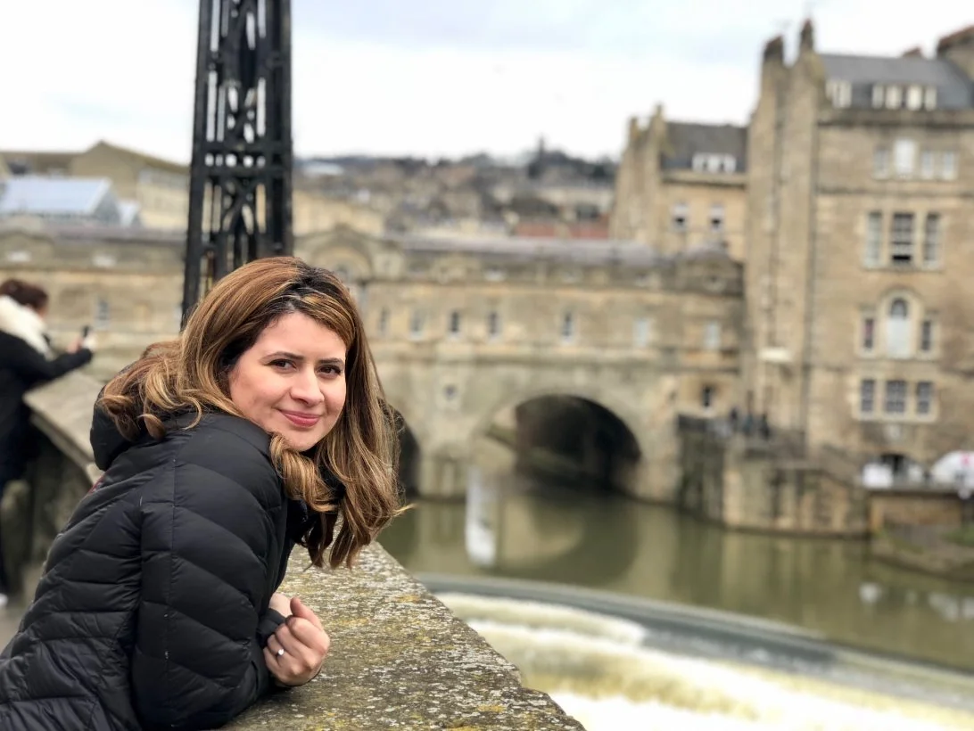A woman leaning on a stone railing along a river, with historic stone buildings in the background.