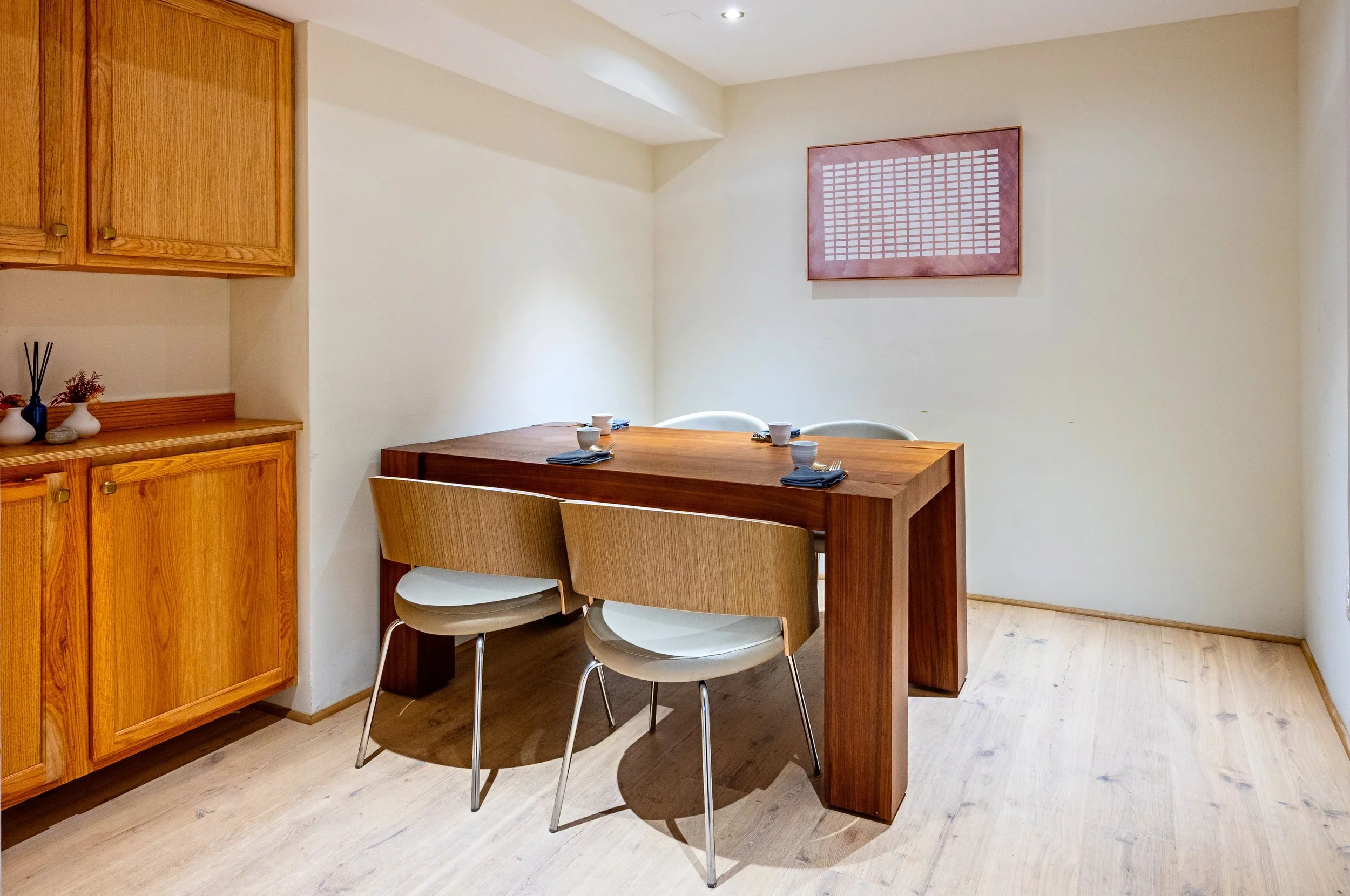 A small dining area with a wooden table set with four cups and napkins, two chairs, and wooden cabinets on the left, with a simple wall decoration.