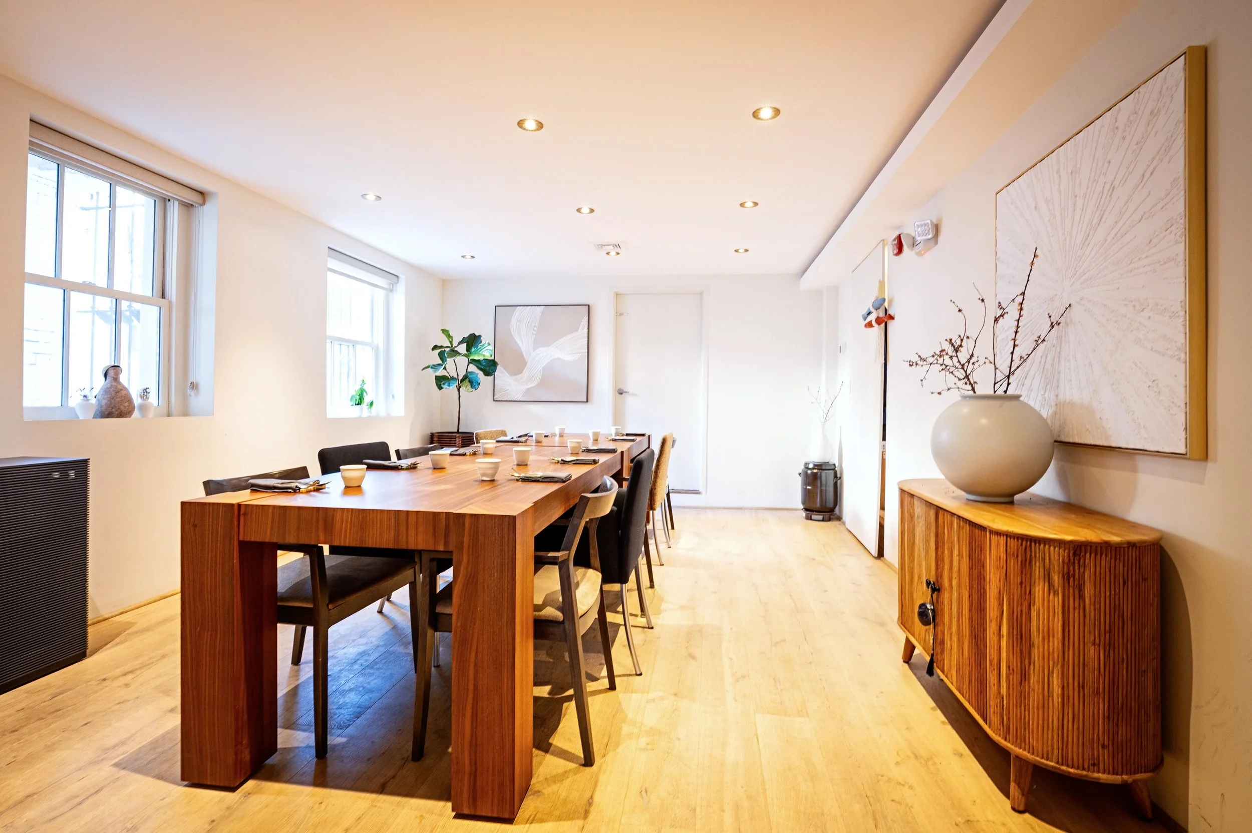 Modern dining room with a large wooden table set for a meal, six black and beige chairs, white walls, abstract artwork, and minimalist decor including potted plants and a vase with branches.