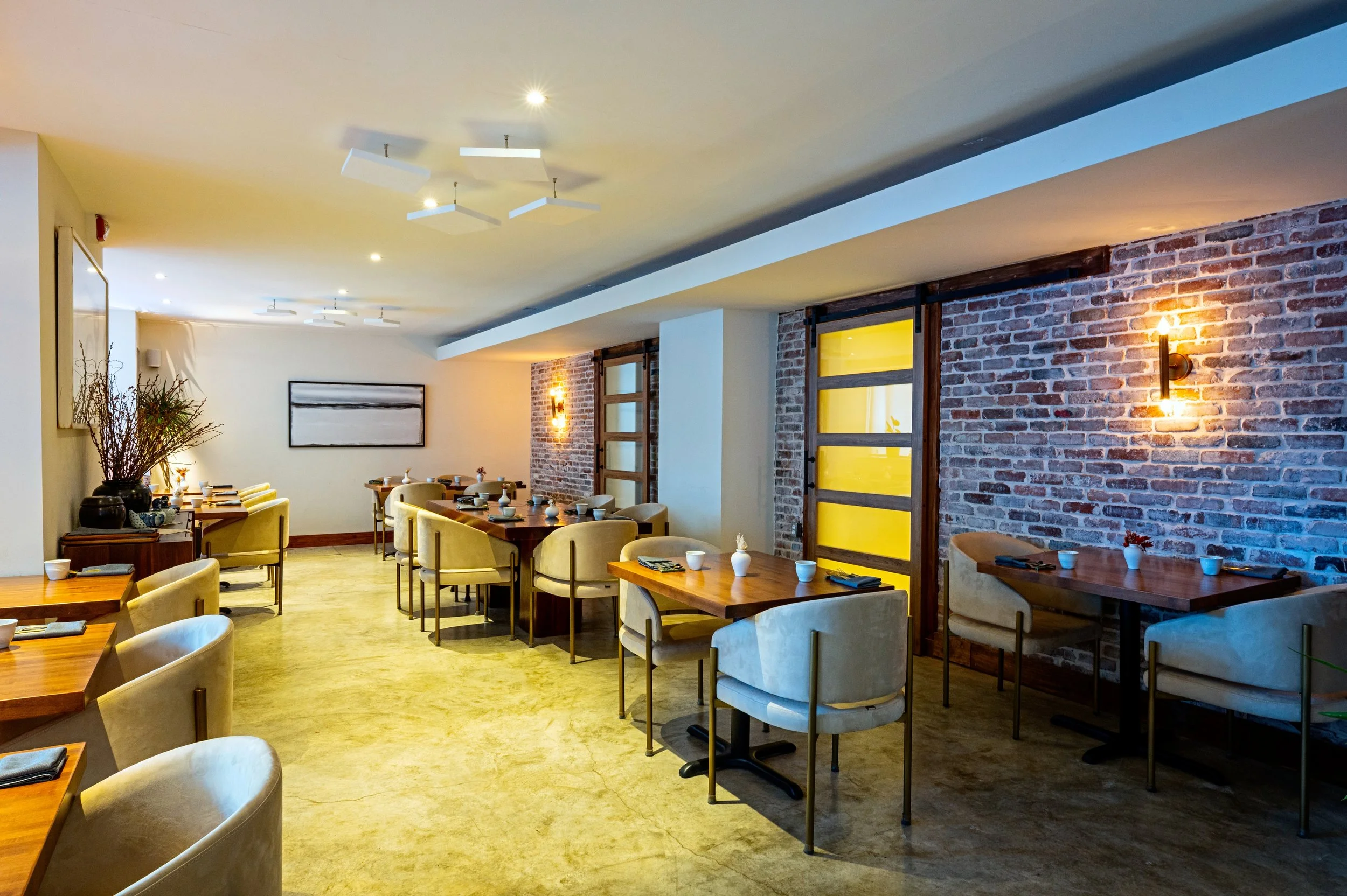Interior of a modern restaurant with wooden tables, yellow chairs, brick walls, and warm lighting.