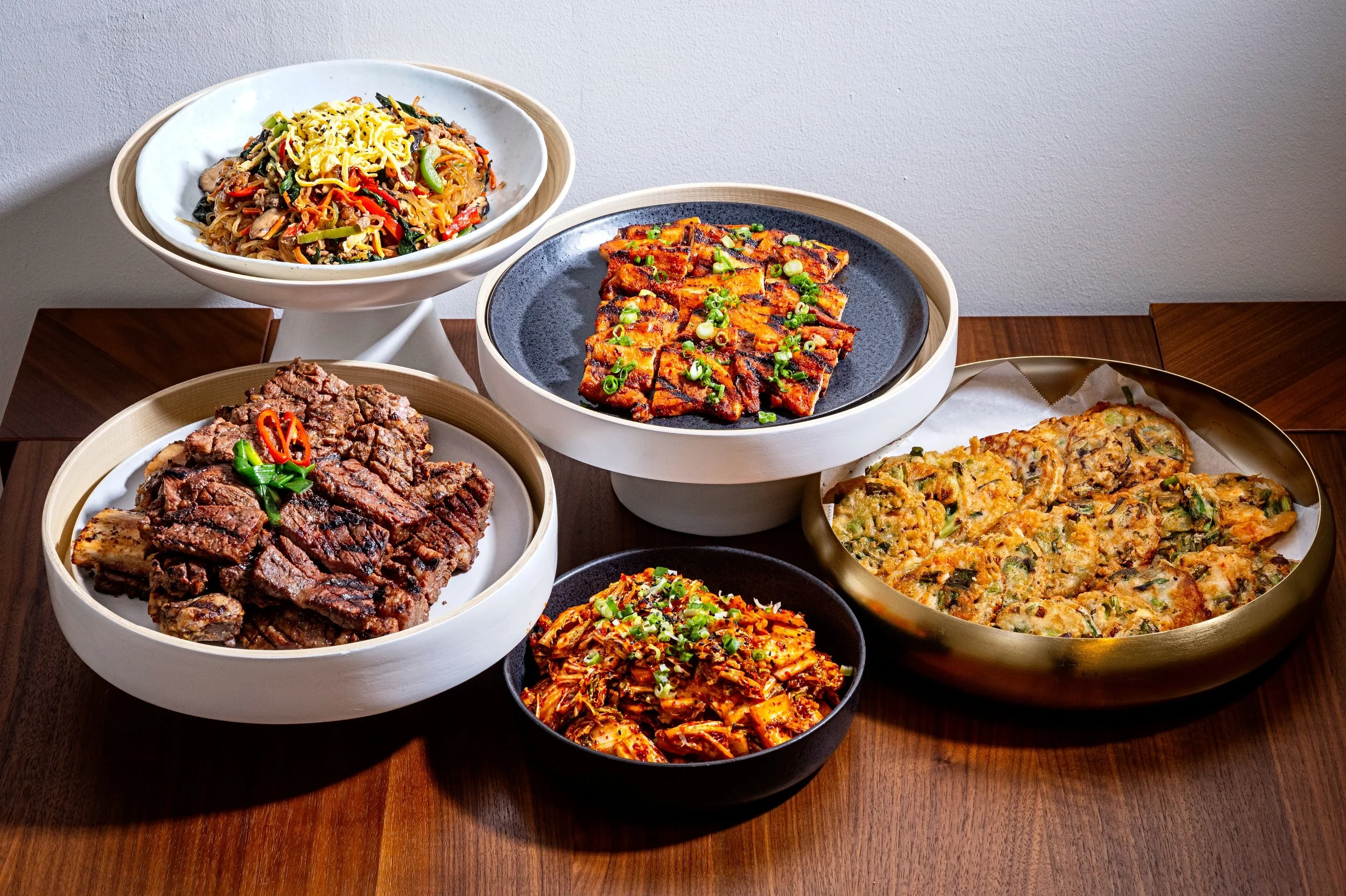 A table with various Korean dishes including stir-fried noodles, grilled pork, kimchi, tofu, and vegetable pancake, all served in bowls and plates.