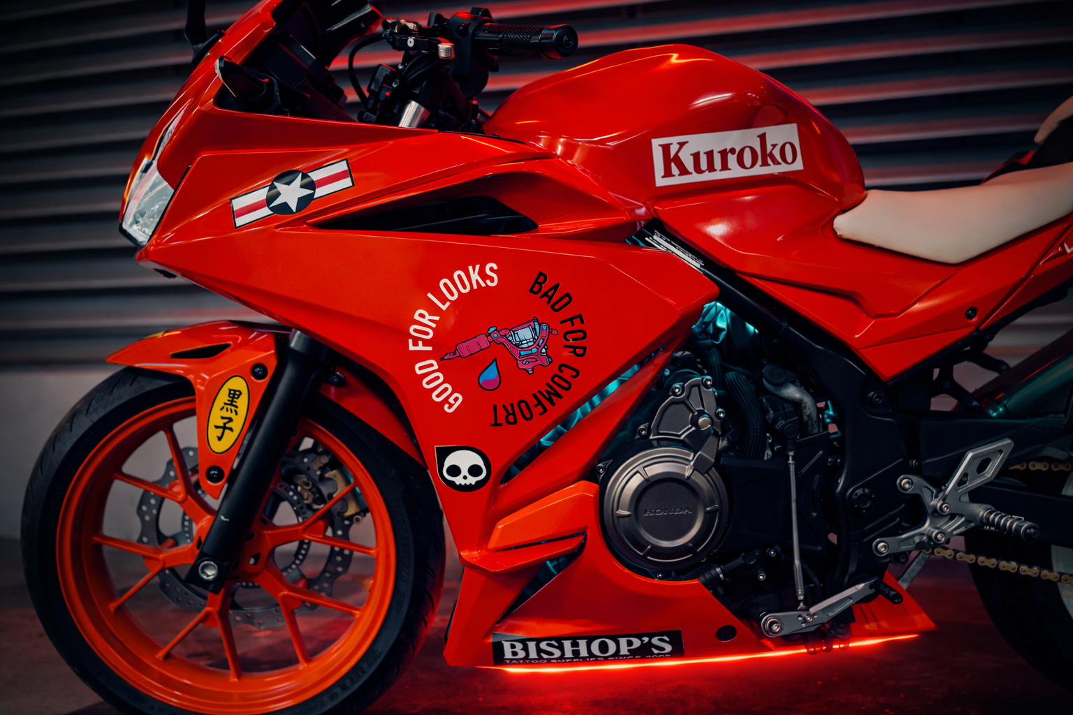 Close-up of a bright orange racing motorcycle with decals and stickers, including a skull symbol, Japanese characters, and slogans like 'Good For Looks, Bad For Comfort,' against a blurred metallic background.