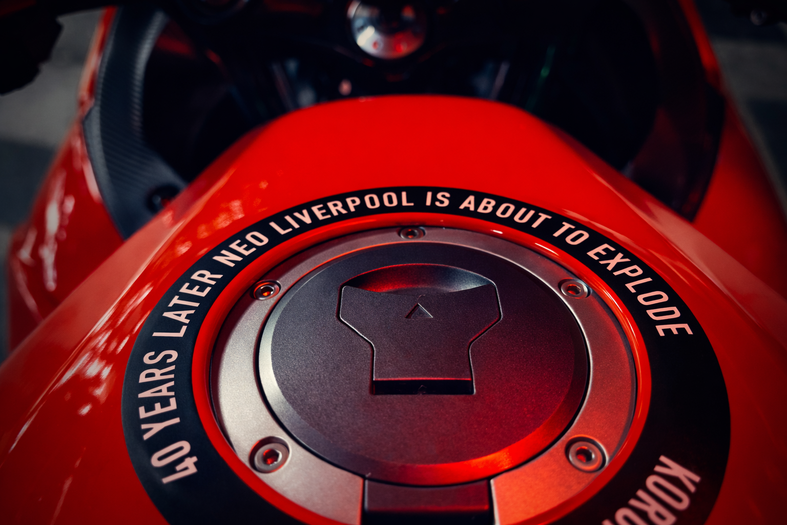 Close-up of a red motorcycle fuel tank with a black cap and a warning sticker about Liverpool.