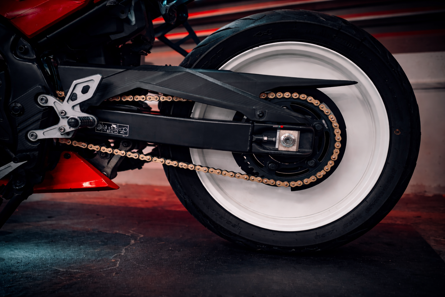 Close-up of a modern motorcycle rear wheel with white rim and gold chain, black swingarm, and tire, set against a dark floor with red and blue lighting reflections.