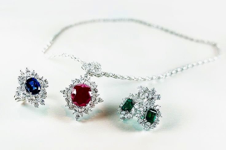 Elegant silver jewelry set with a ruby pendant necklace, a sapphire ring, and emerald earrings with diamond accents on a light background.