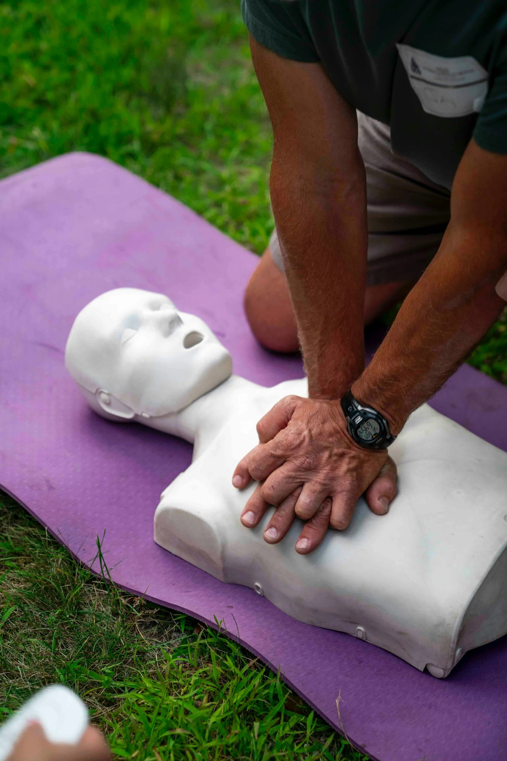 Person performing CPR on a mannequin outdoors on a purple mat.