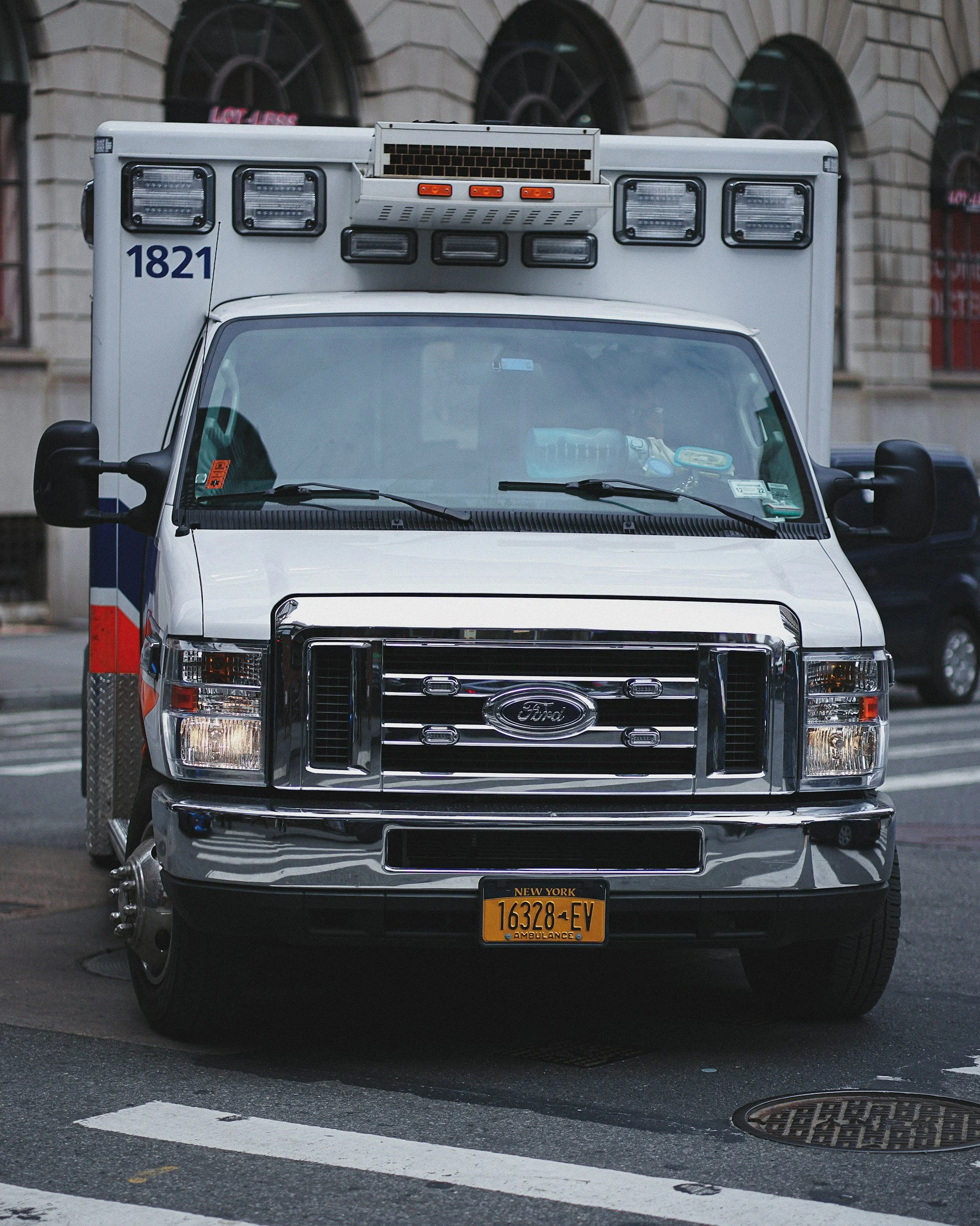Front view of a white ambulance vehicle parked on a street in New York. The vehicle has emergency lights, a front grille, and a New York license plate reading 16328-FV. In the background, there is a building with arched windows.