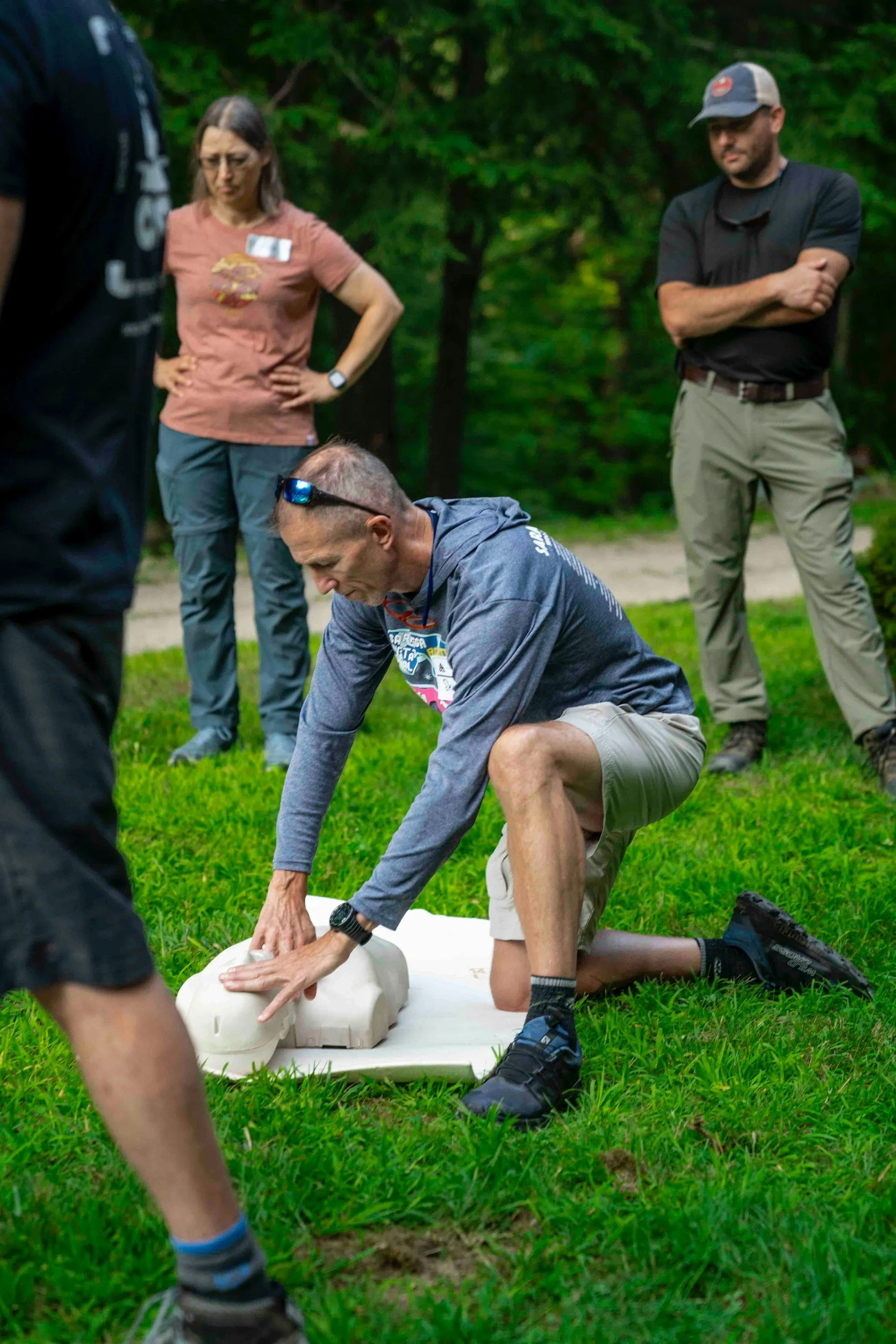 Group of people participating in a CPR training class outdoors, practicing chest compressions on a dummy on green grass, with trees in the background.