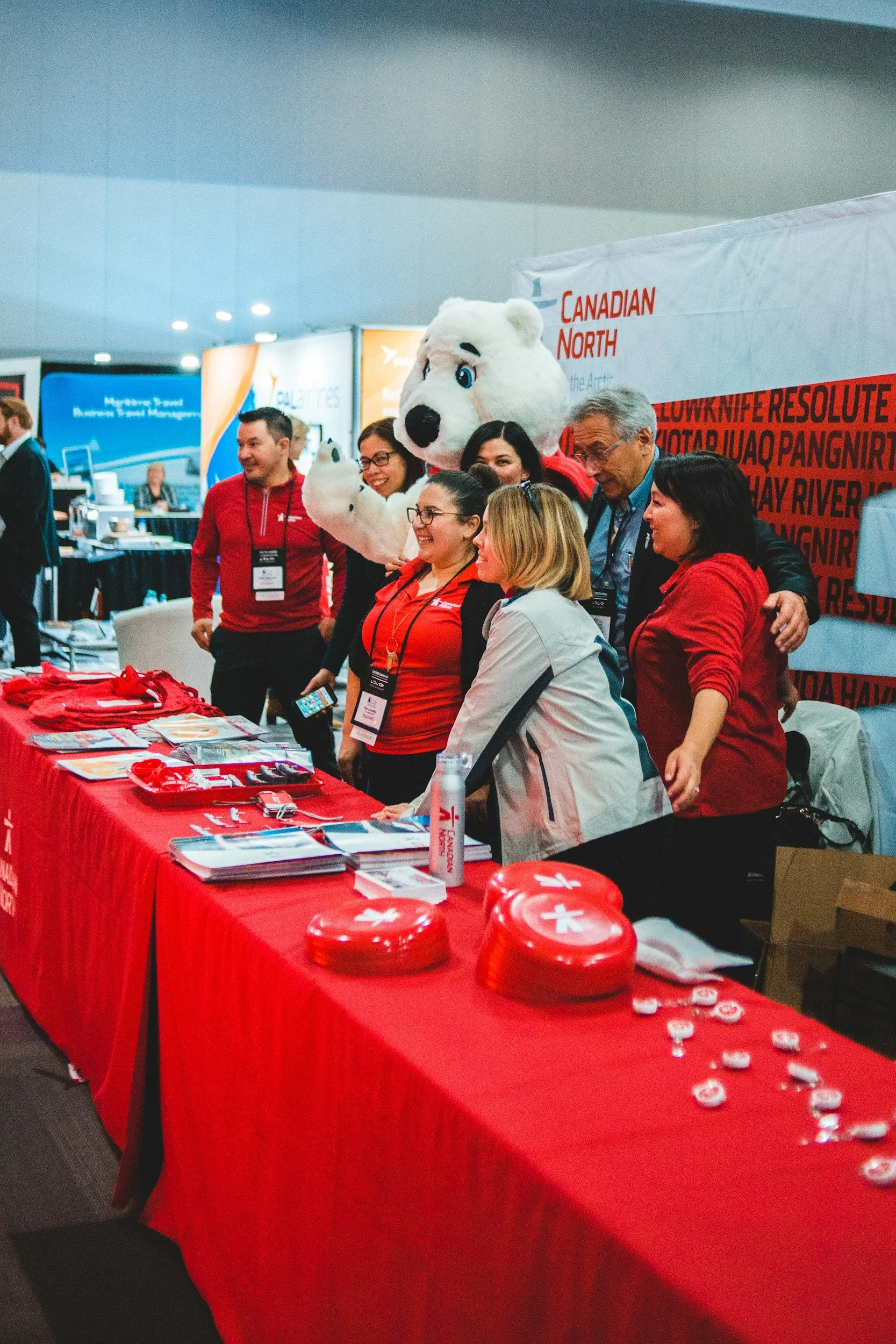 A group of people, including a person in a polar bear costume, standing at a booth with a red tablecloth. The booth has brochures, round red containers with white crosses, and white buttons. The background has banners with flight information and the words "Canadian North."