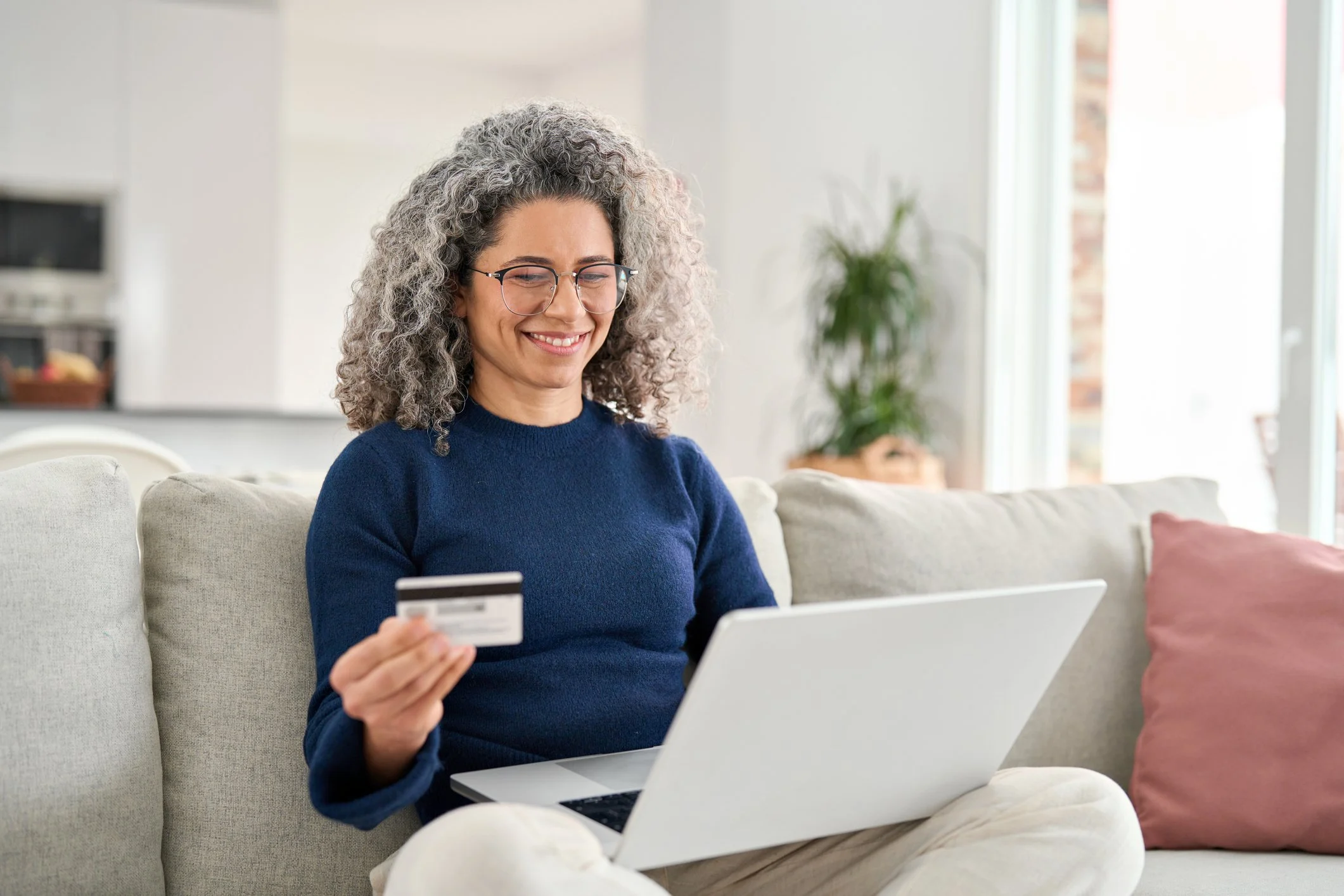 Woman sitting on a beige sofa with a laptop on her lap, holding a credit card in her hand, smiling at the screen.
