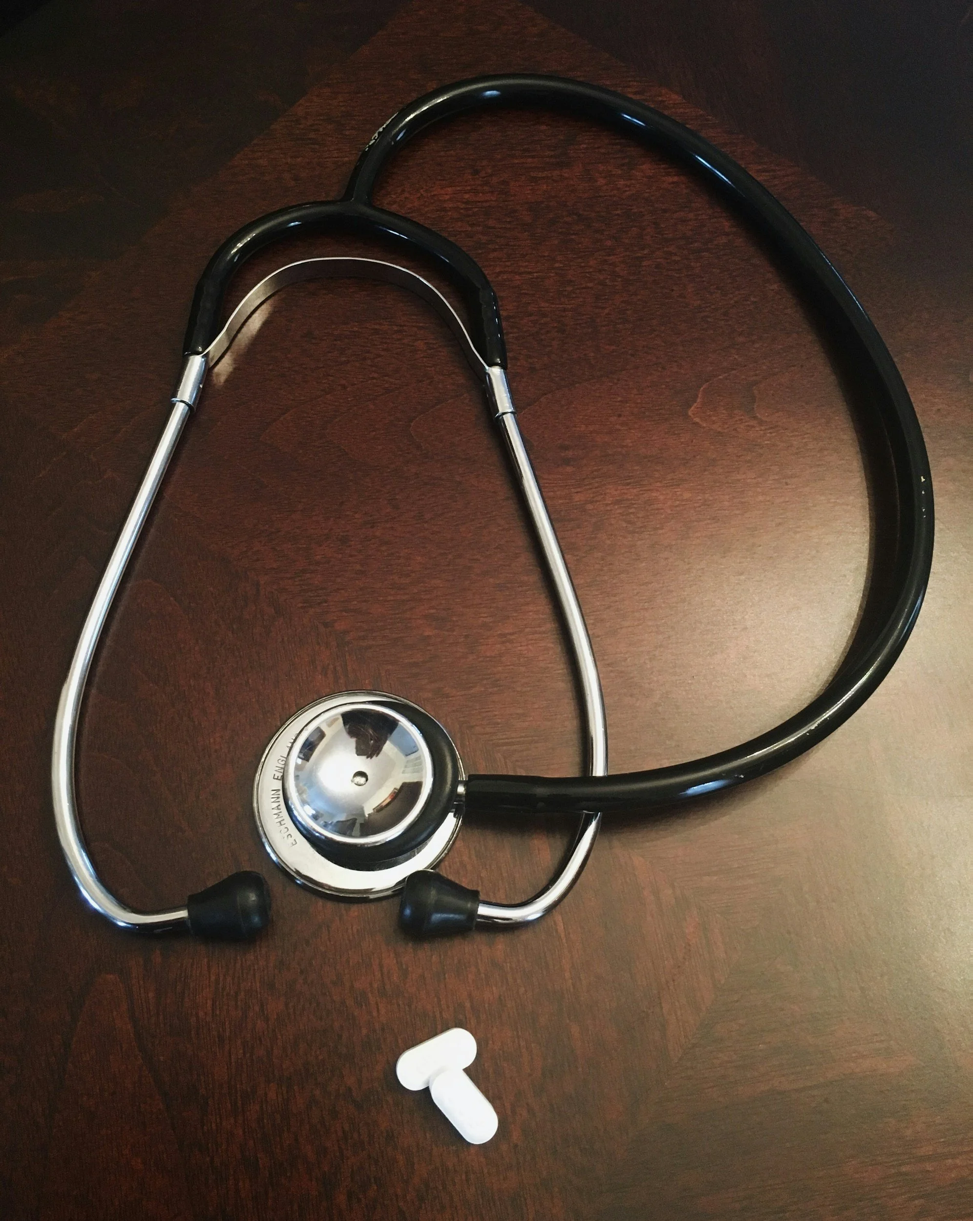A black and silver stethoscope with black ear tips and a white pill on a wooden surface.