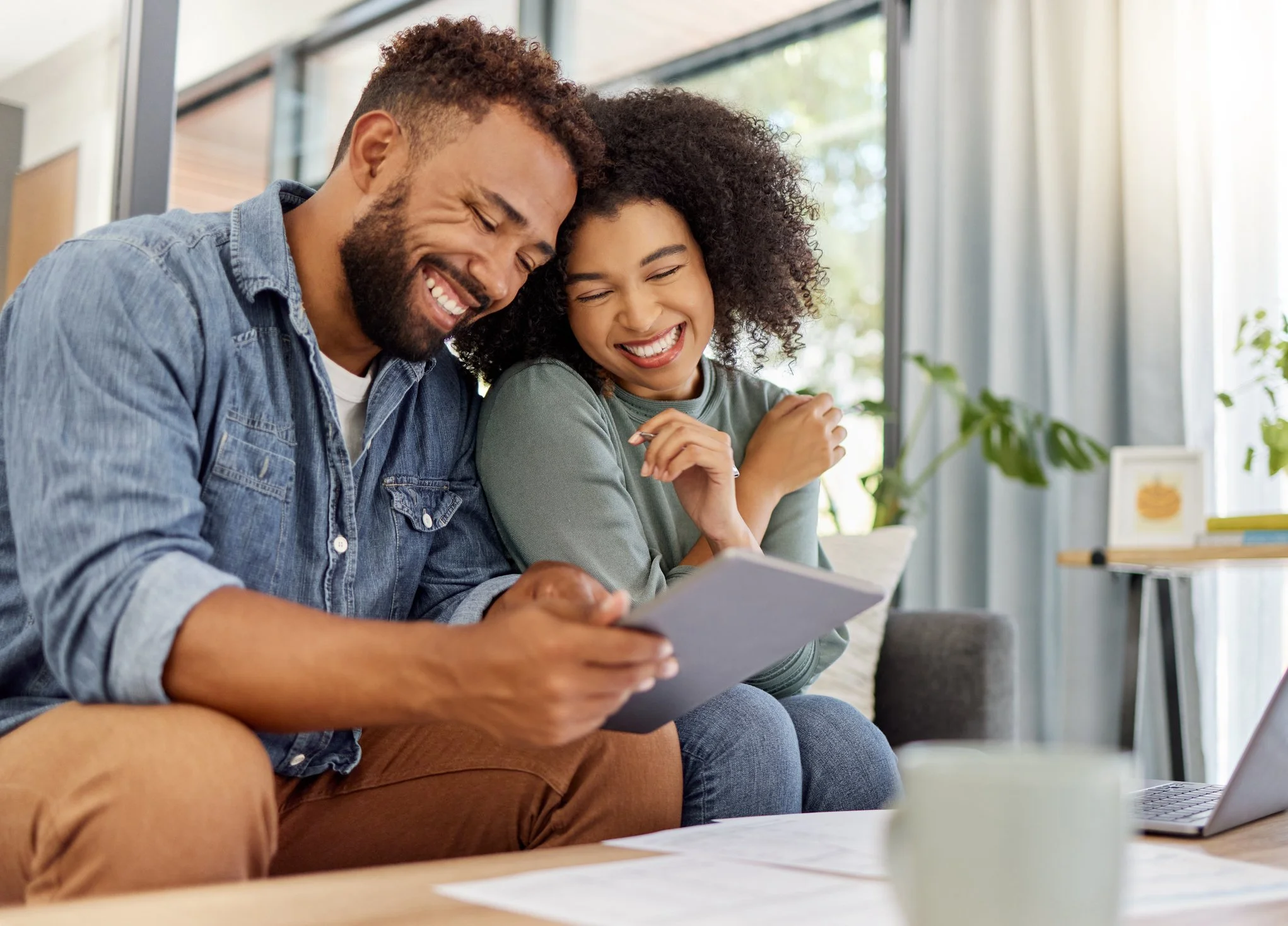Smiling man and woman sitting together, looking at a tablet in a bright living room.
