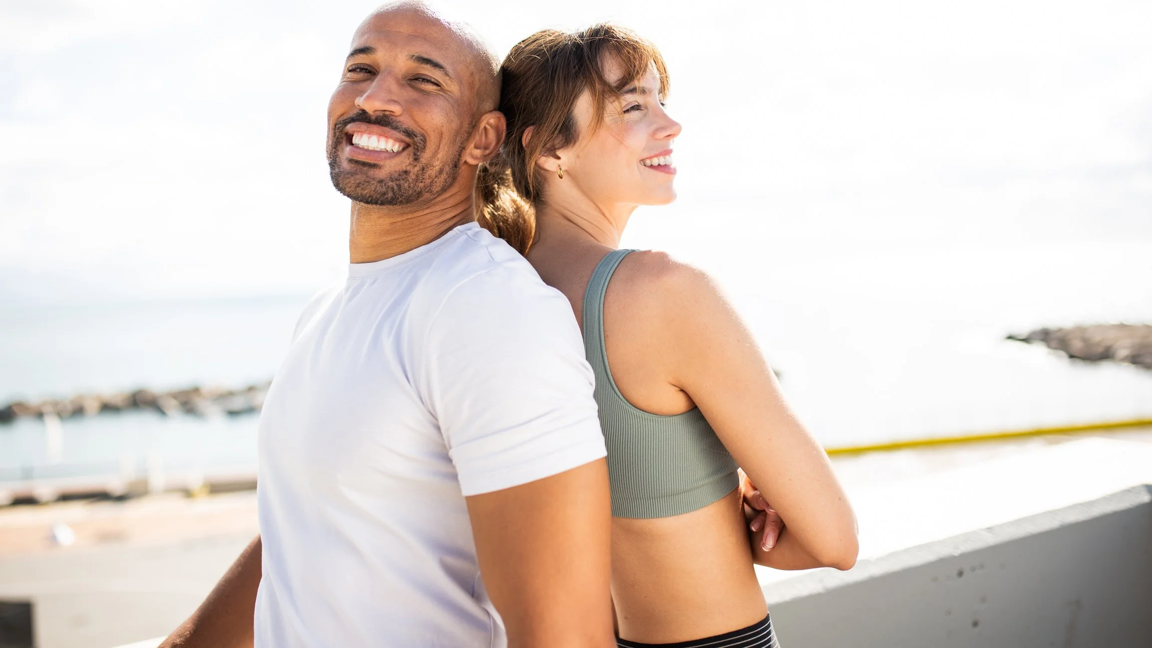 A smiling man and woman stand back to back outdoors near a body of water on a sunny day, with relaxed expressions and casual clothing.