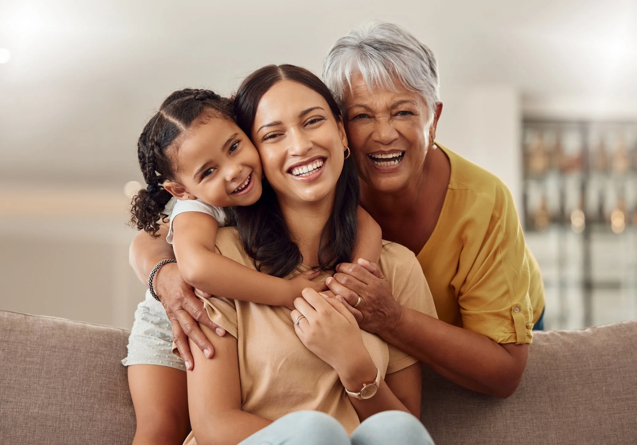Three generations of women smiling and hugging on a beige couch in a bright living room.