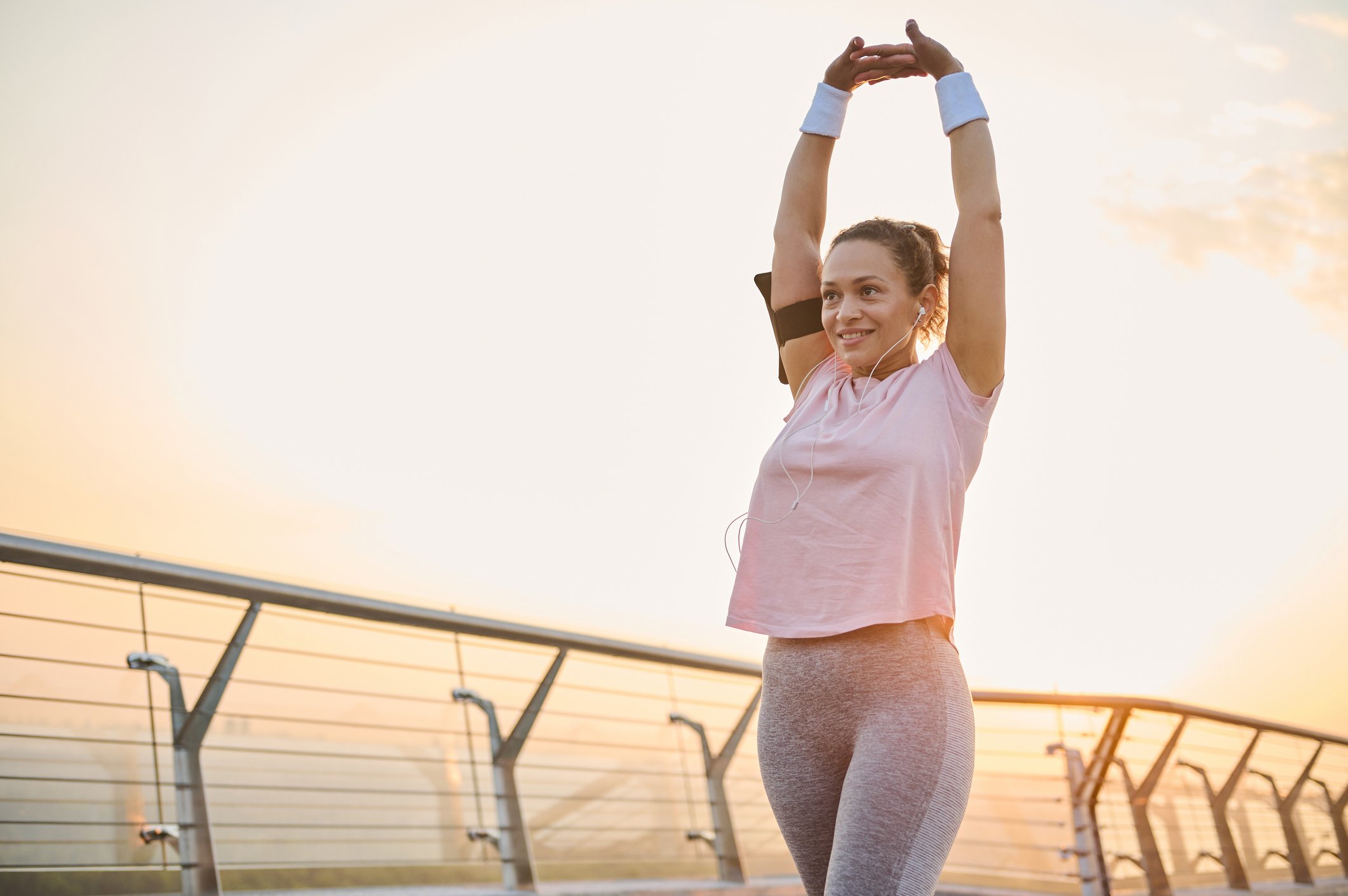 A young woman stretching outdoors during sunset, wearing workout clothes and earphones.