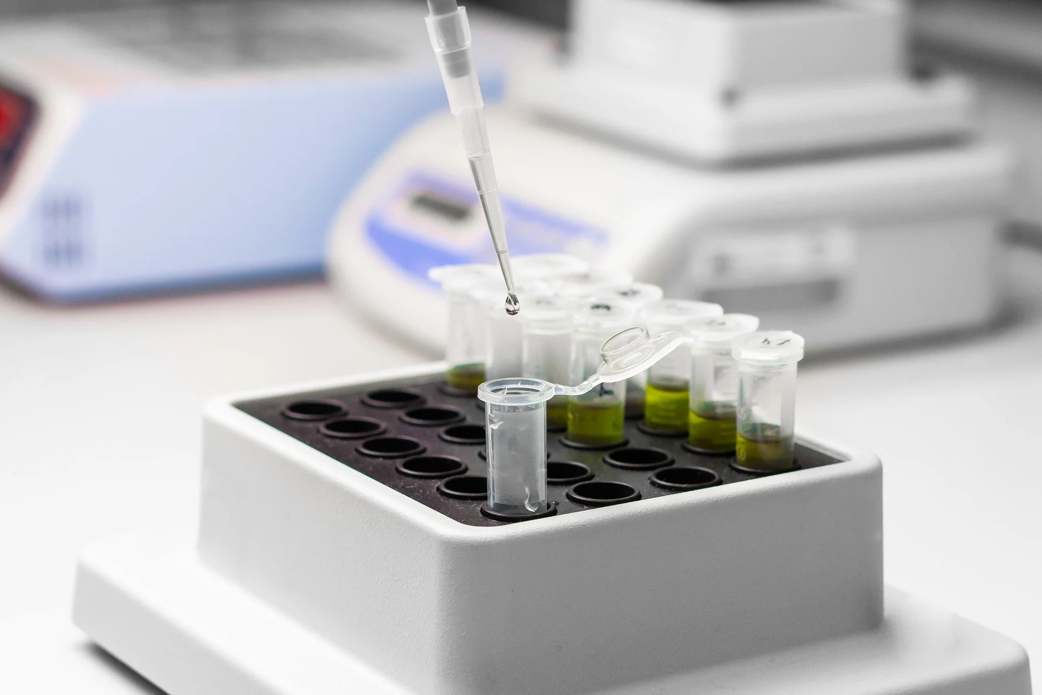 Laboratory scene with a pipette dripping liquid into a test tube rack holding several test tubes with green liquid.