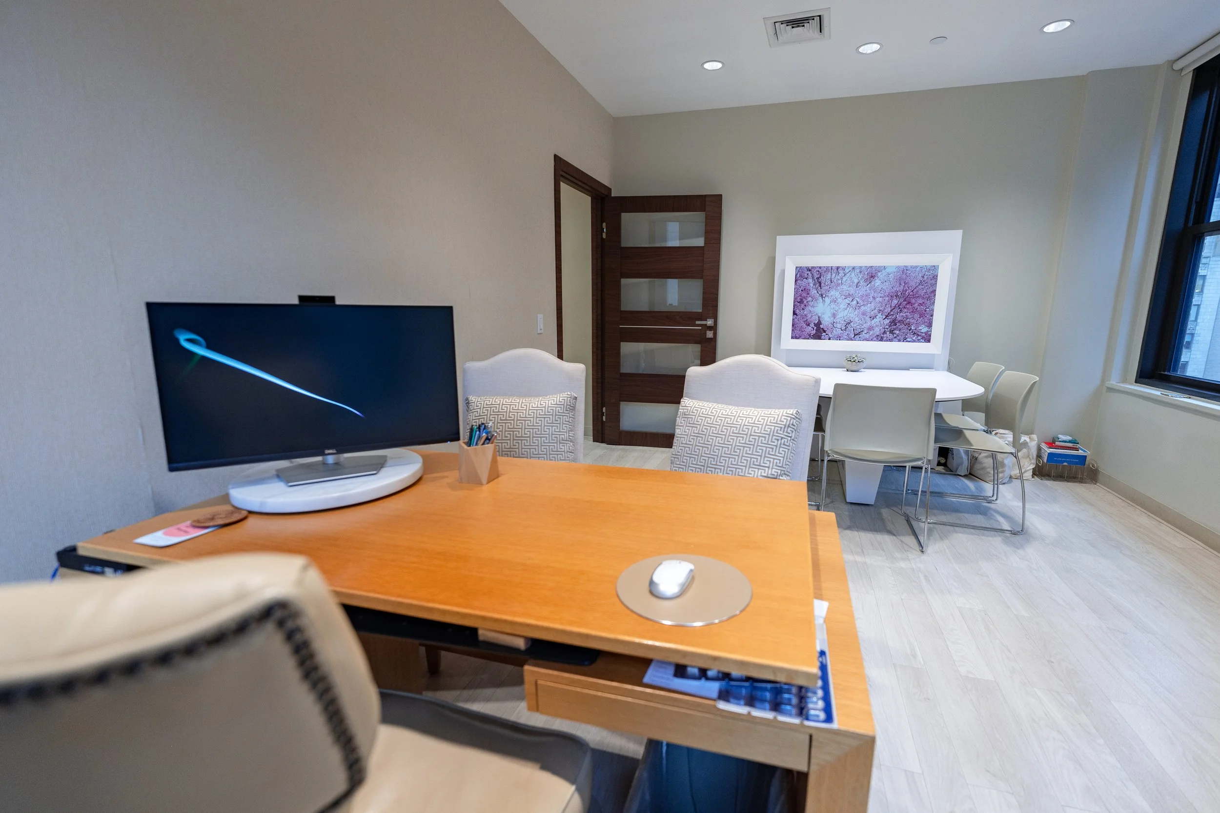 Office room with a wooden desk, a computer monitor, a mouse, a chair, and a table with chairs in the background. There is a window with a view outside and some boxes on the floor.