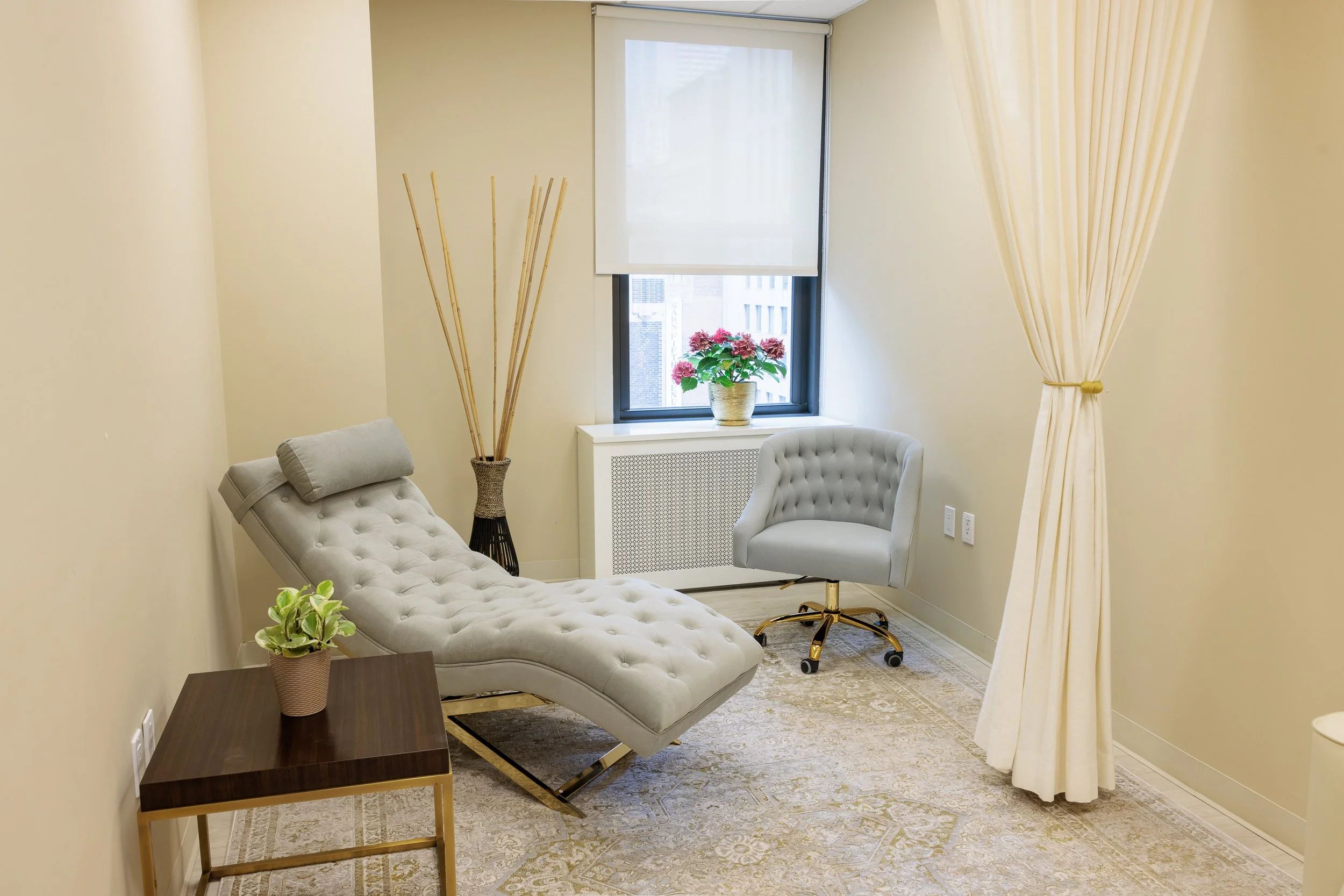 Modern beige office or lounge room with tufted chaise lounge, gray swivel chair, wooden side table with potted plant, window with white shade, potted pink flowers on radiator cover, and beige curtains.