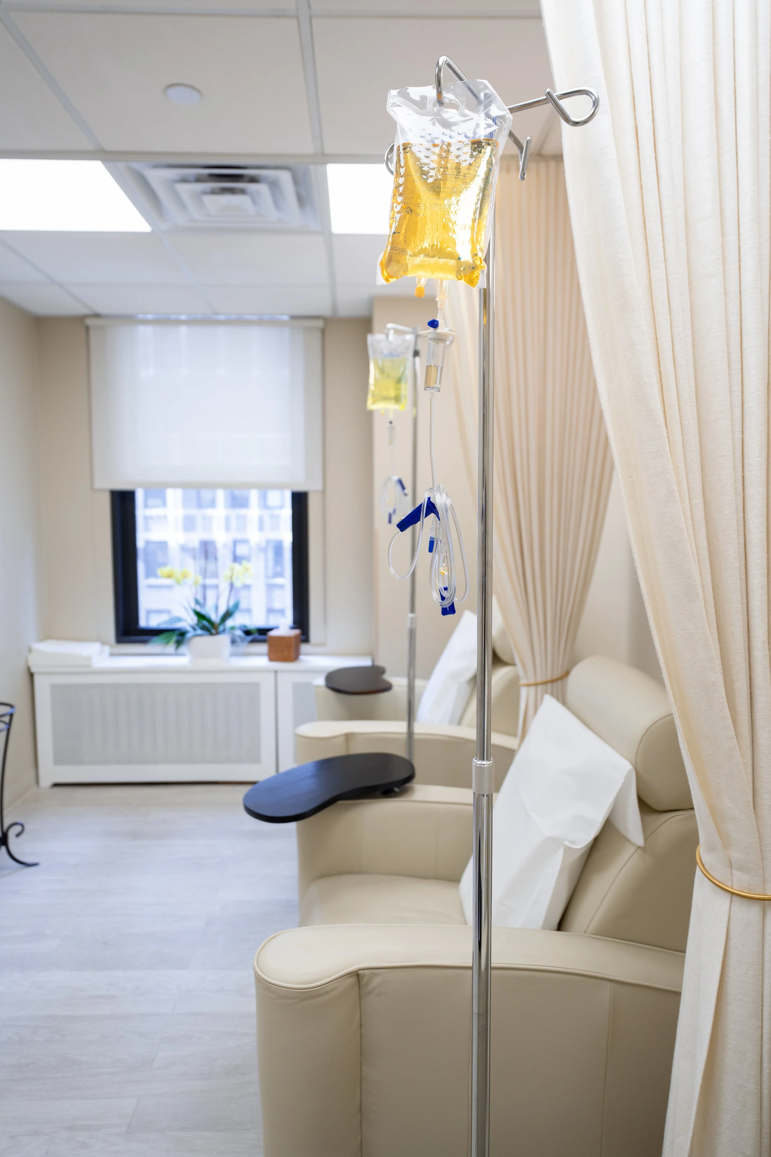 Hospital room with IV poles and bags of IV fluids, beige recliner chairs, and a window with a plant on the windowsill.