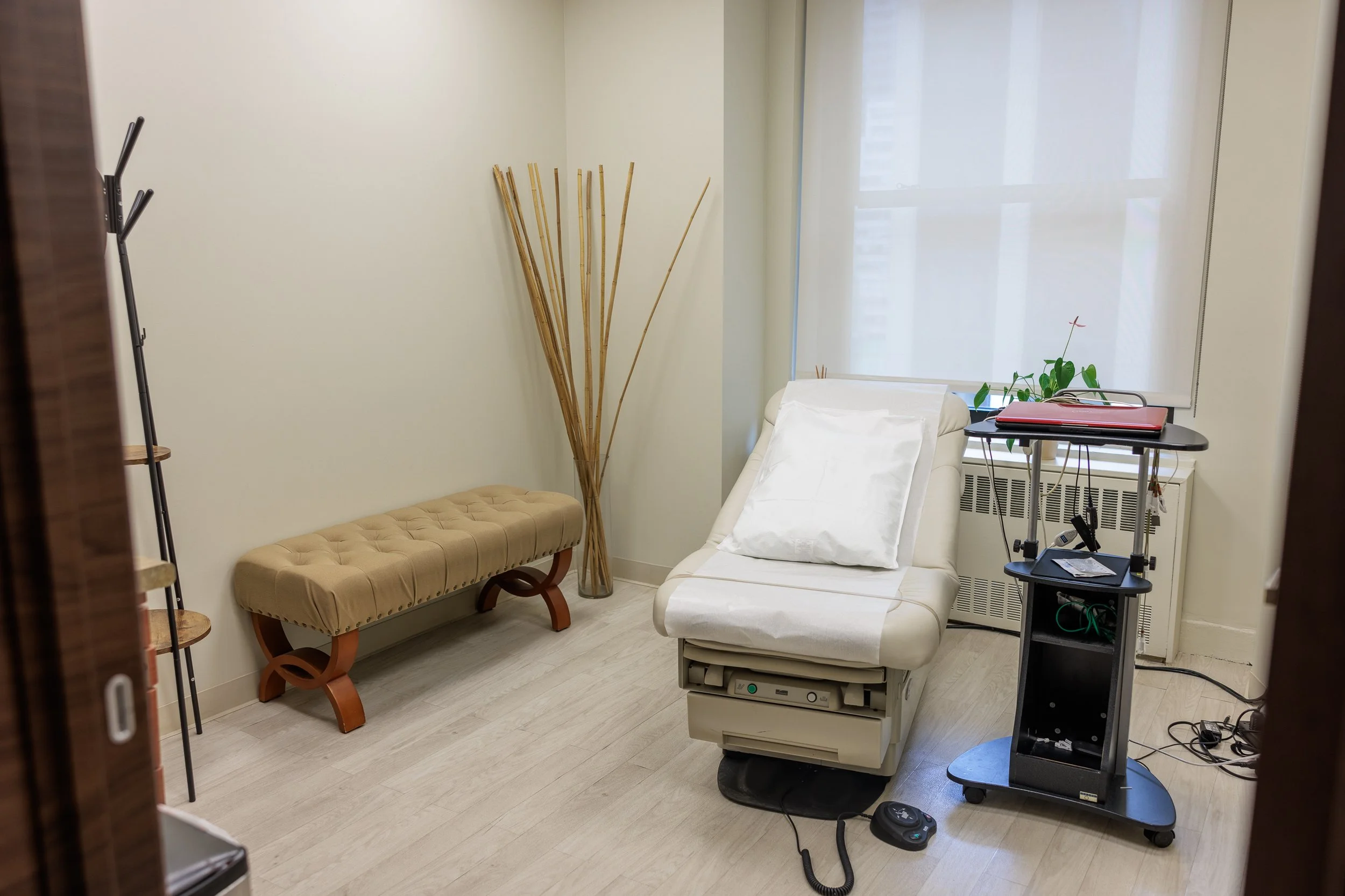 Medical examination room with a reclining bed, a beige bench, a tall glass vase with decorative sticks, a window with blinds, and medical equipment on a stand.