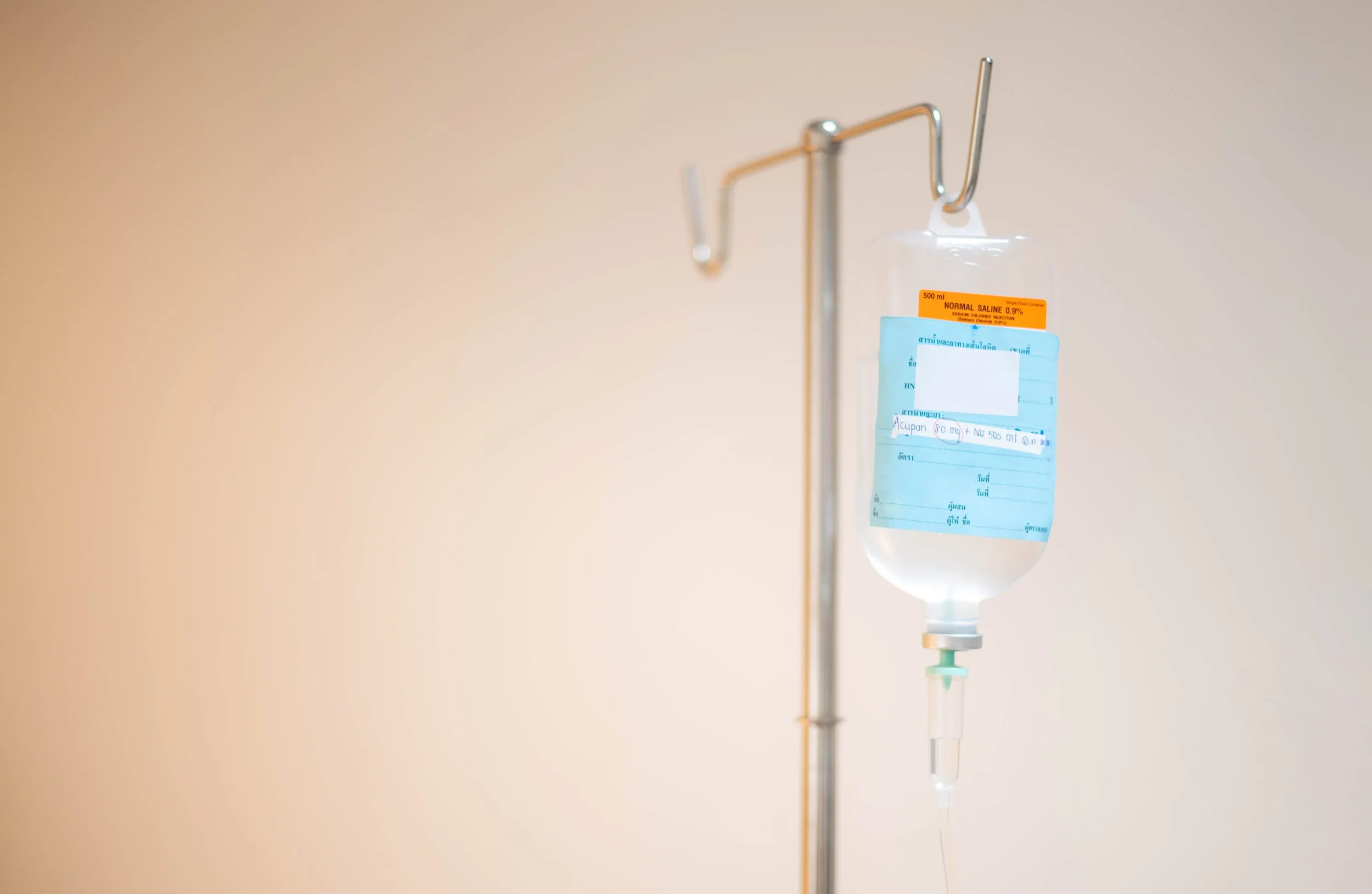 Hospital IV drip bag filled with clear liquid hanging from an adjustable stand against a neutral background.