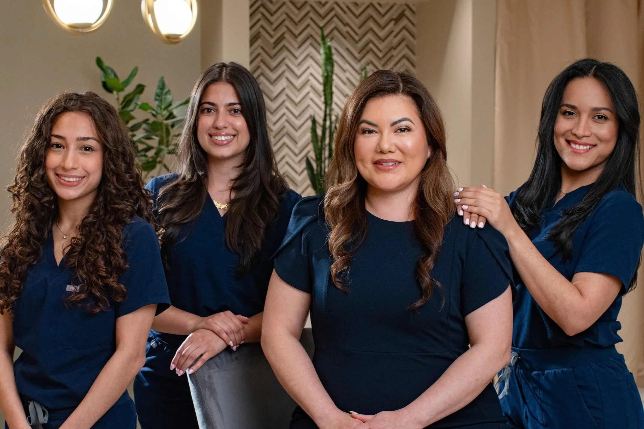 Four women in navy blue scrubs posing together in a modern setting with plants and decorative wall patterns.