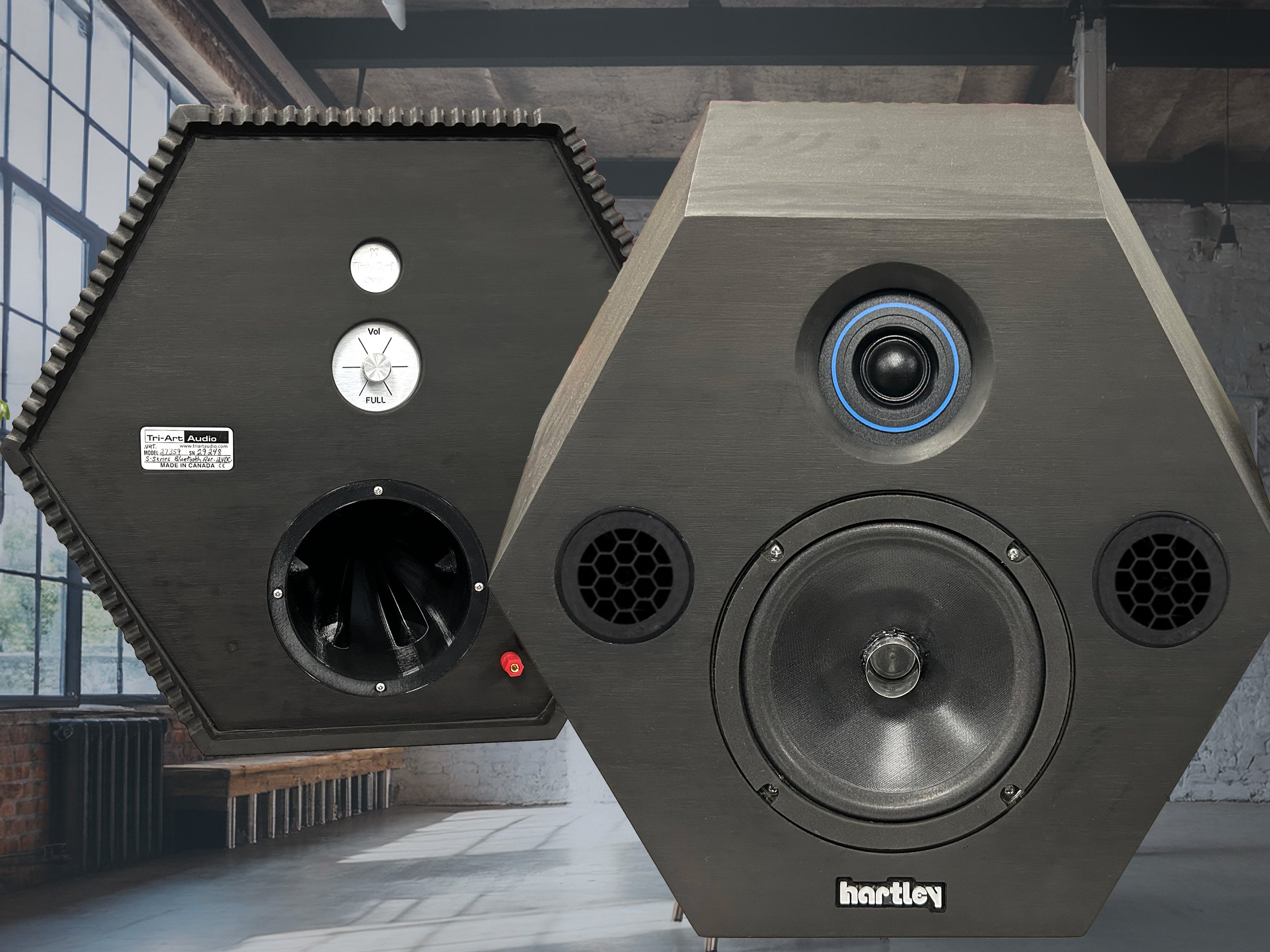 Two large black speakers with hexagonal design, set against an industrial-style interior with large windows and exposed brick and concrete walls.