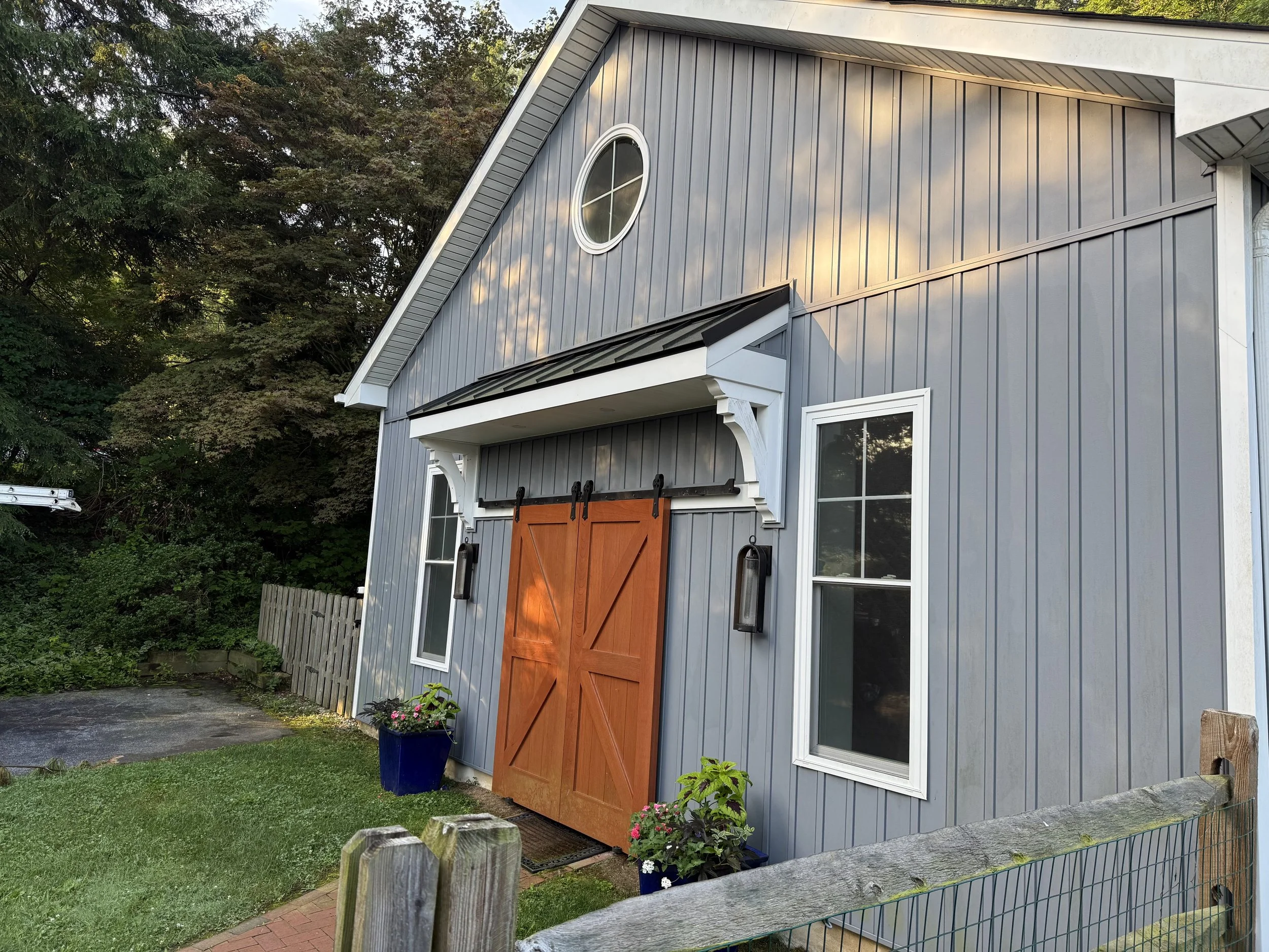 A gray metal building with a pitched roof and a circular window at the top. It has two white-framed windows and a wooden sliding barn door with black hardware. Two outdoor wall lanterns are mounted on either side of the door, with a blue flower pot a