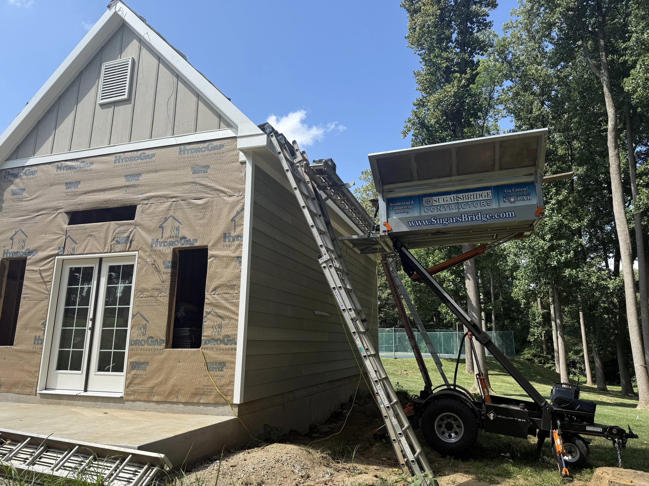 Construction site of a house with siding being installed, ladder leaning on the house, and a lift with the contractor company's sign.