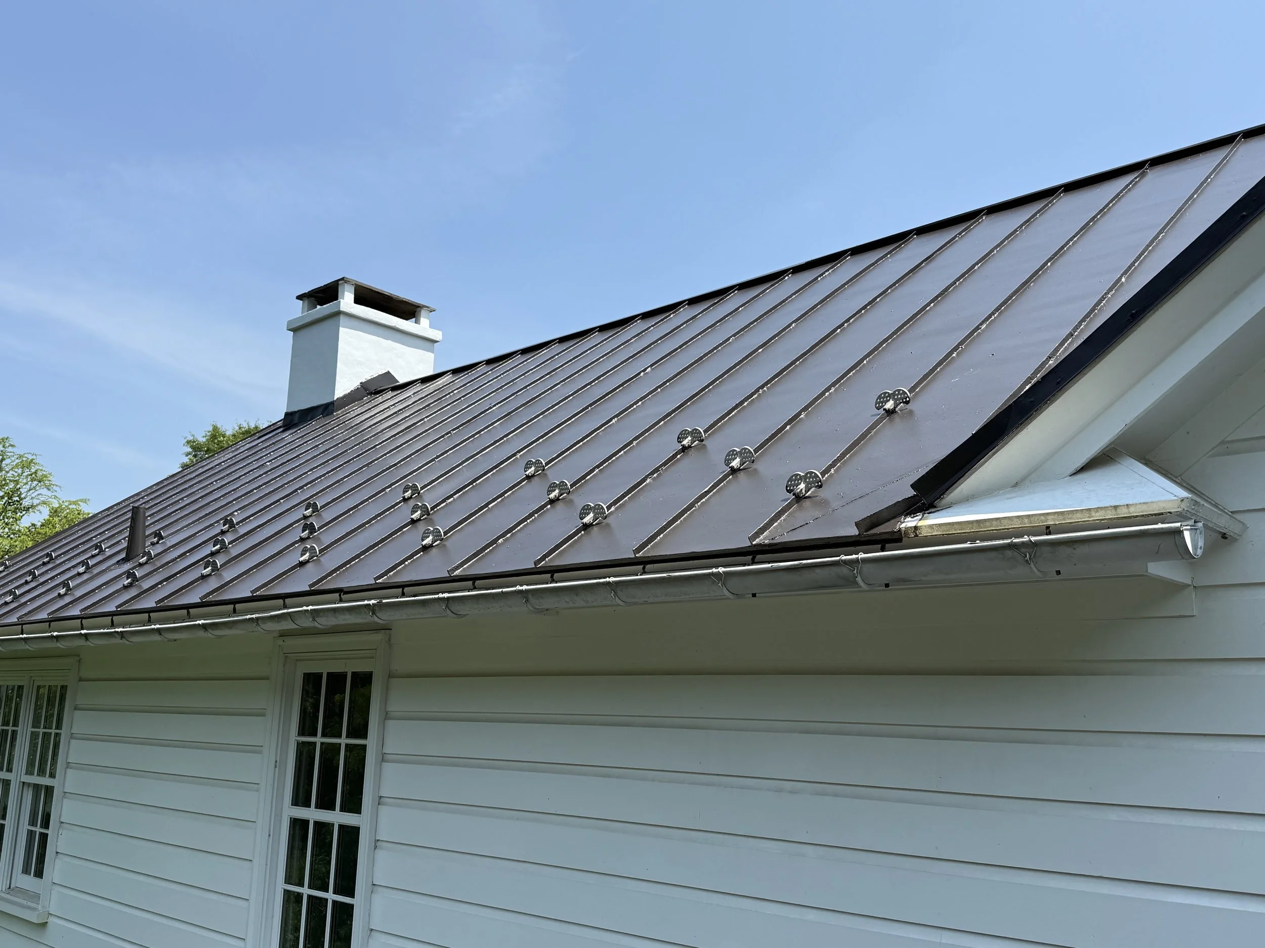 House roof with metal roofing panels and snow guards, white siding, two windows, and a chimney with a cap under a clear blue sky.