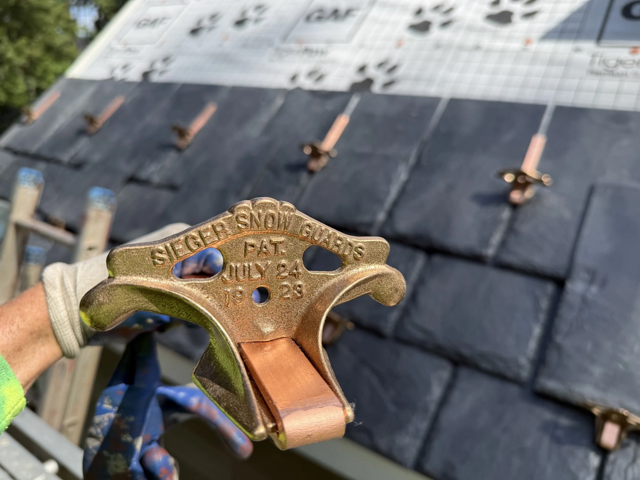A person holding a copper roofing nail pulling a slate roof tile in the background.