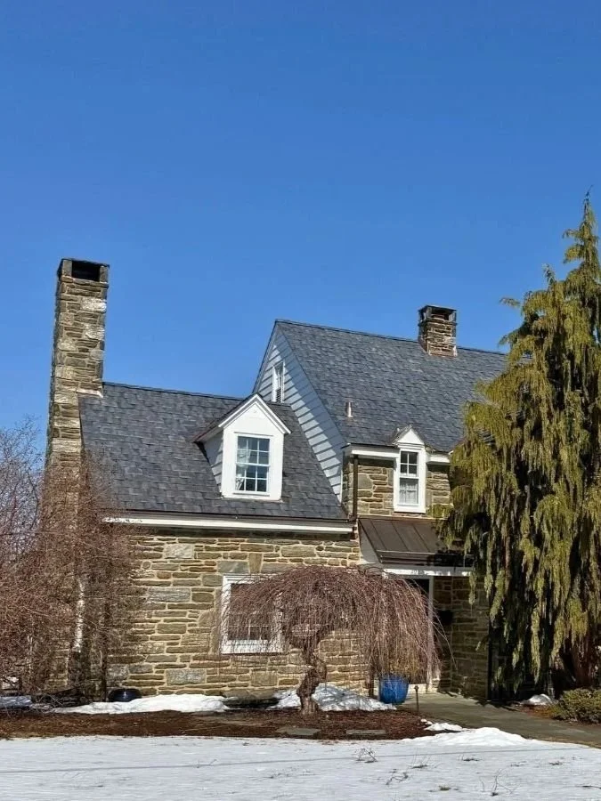 Stone house with gray shingle roof, two chimneys, white dormer windows, and a large tree in front under a clear blue sky.