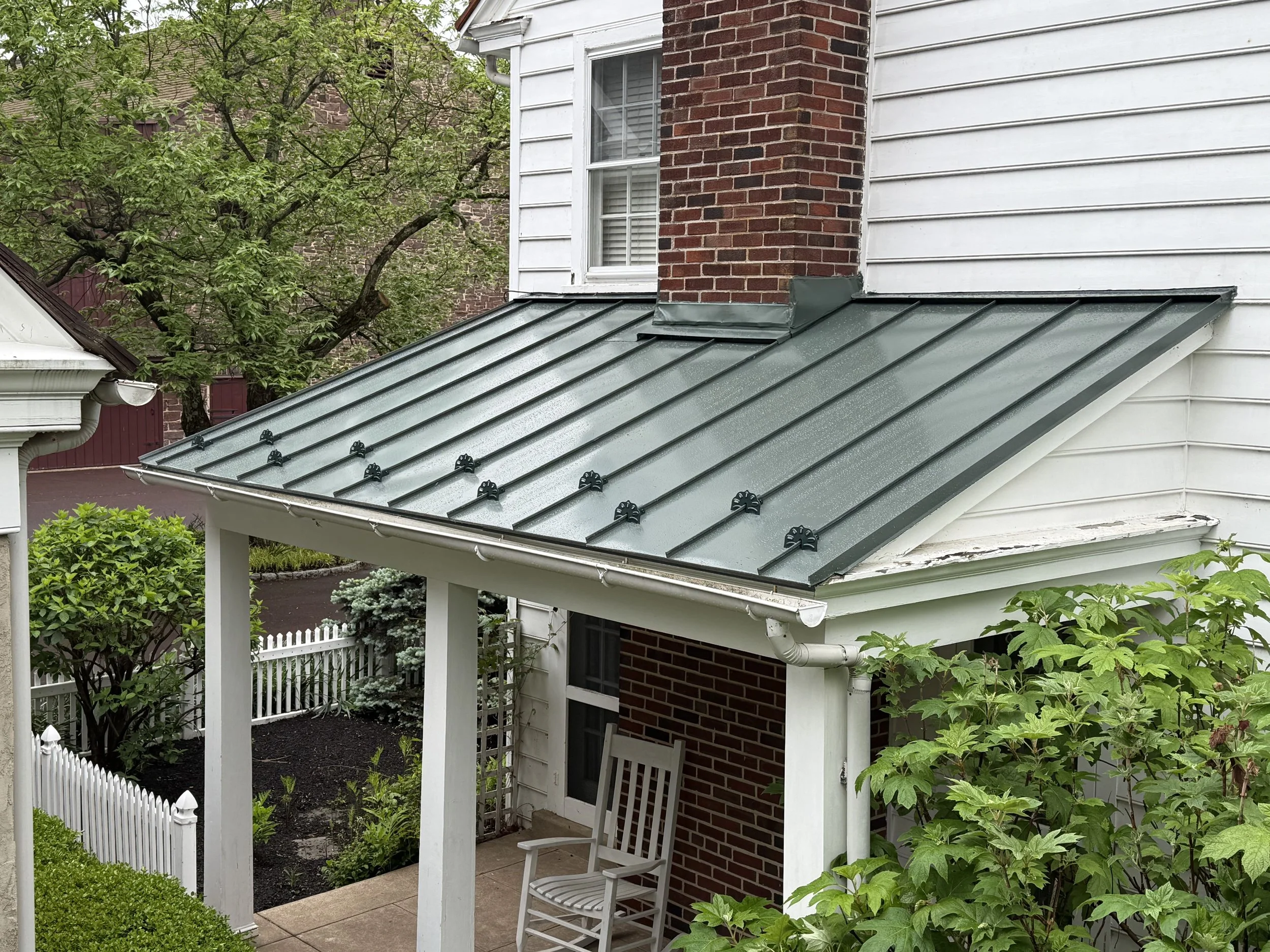 Metal roof with fasteners on the edge of a porch of a house with white siding and brick wall, surrounded by green trees and plants.