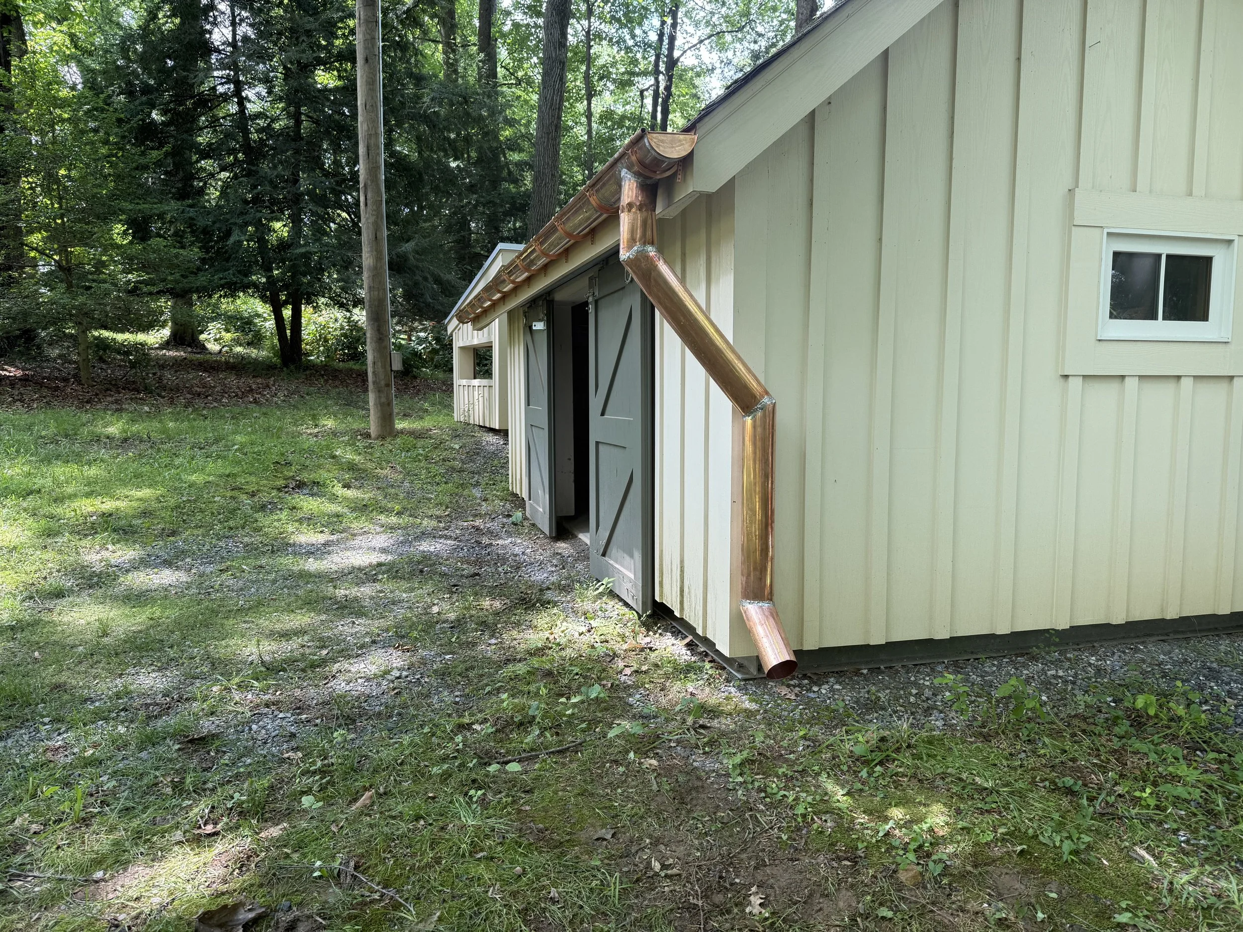 A yellow wooden shed with two gray double doors and a small window. There is a copper gutter system attached to the roof, extending down towards the ground. The shed is on a grassy area with trees and a utility pole in the background.