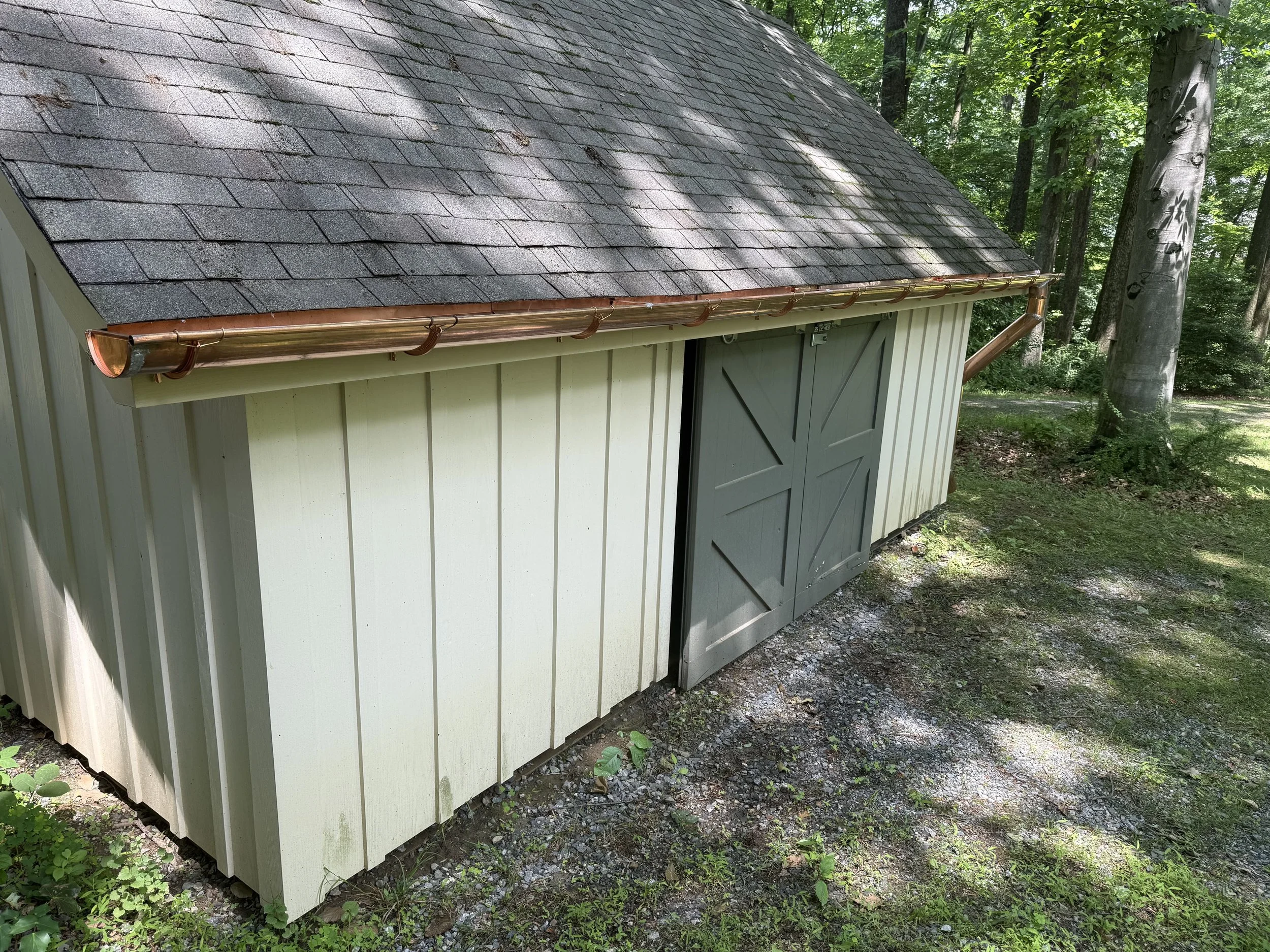 Small shed with a gray double barn door, off-white wooden siding, and a shingled roof, situated in a shaded wooded area with trees and grass.