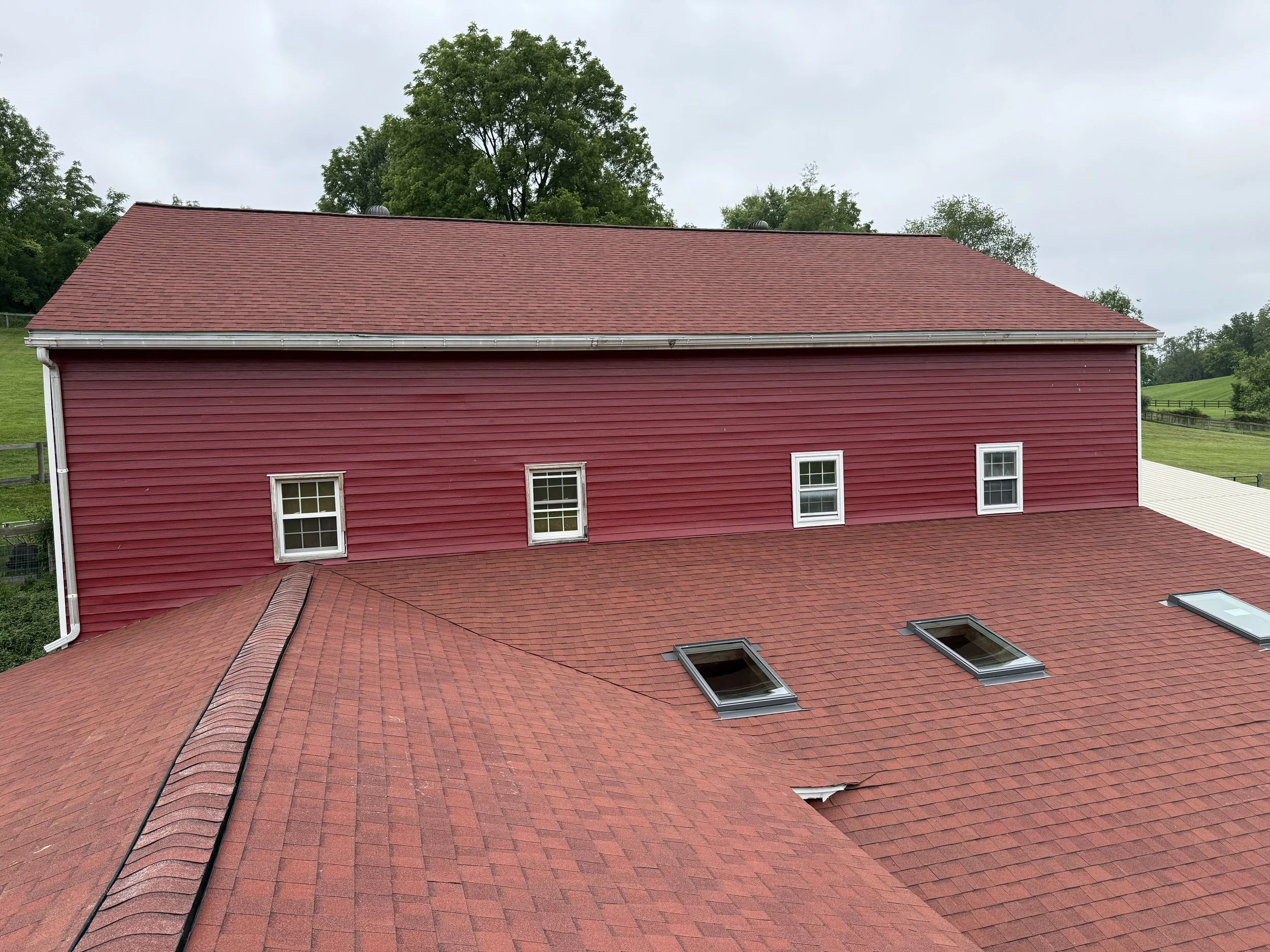 A red barn with three small windows on the upper level and three skylights on the lower roof, set in a rural landscape with trees and grassy fields in the background.