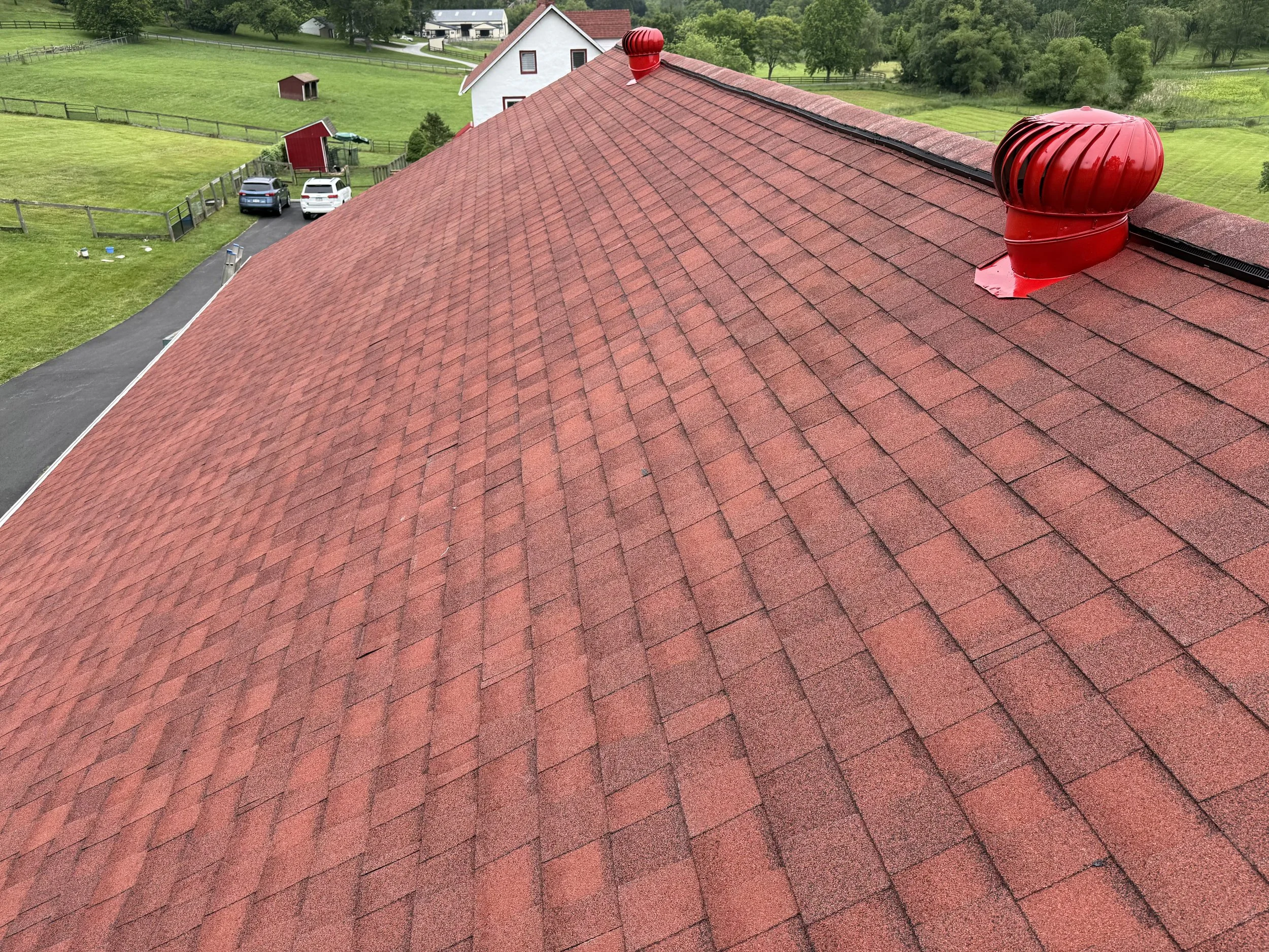 Close-up view of a red shingle roof with two red exhaust vents, overlooking a green countryside with houses, trees, and a driveway with parked cars.
