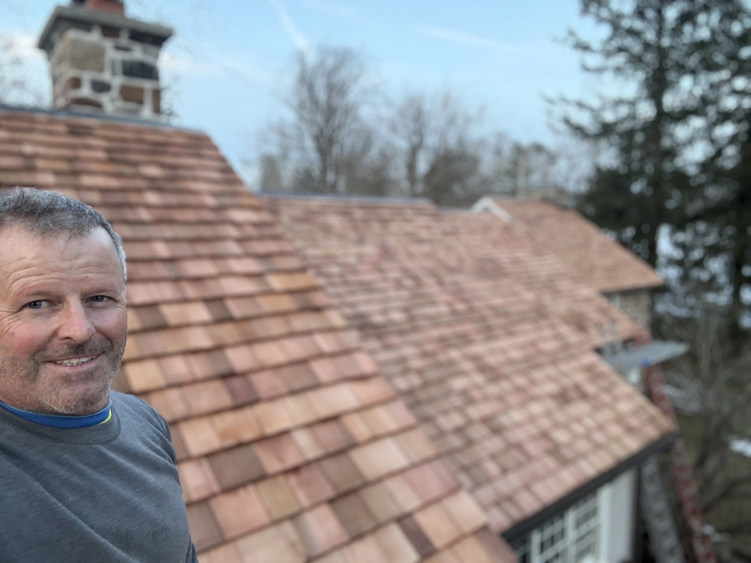 A man with short hair and a beard taking a selfie in front of a house with a tiled roof. The roof has reddish-brown tiles, and there are trees in the background.