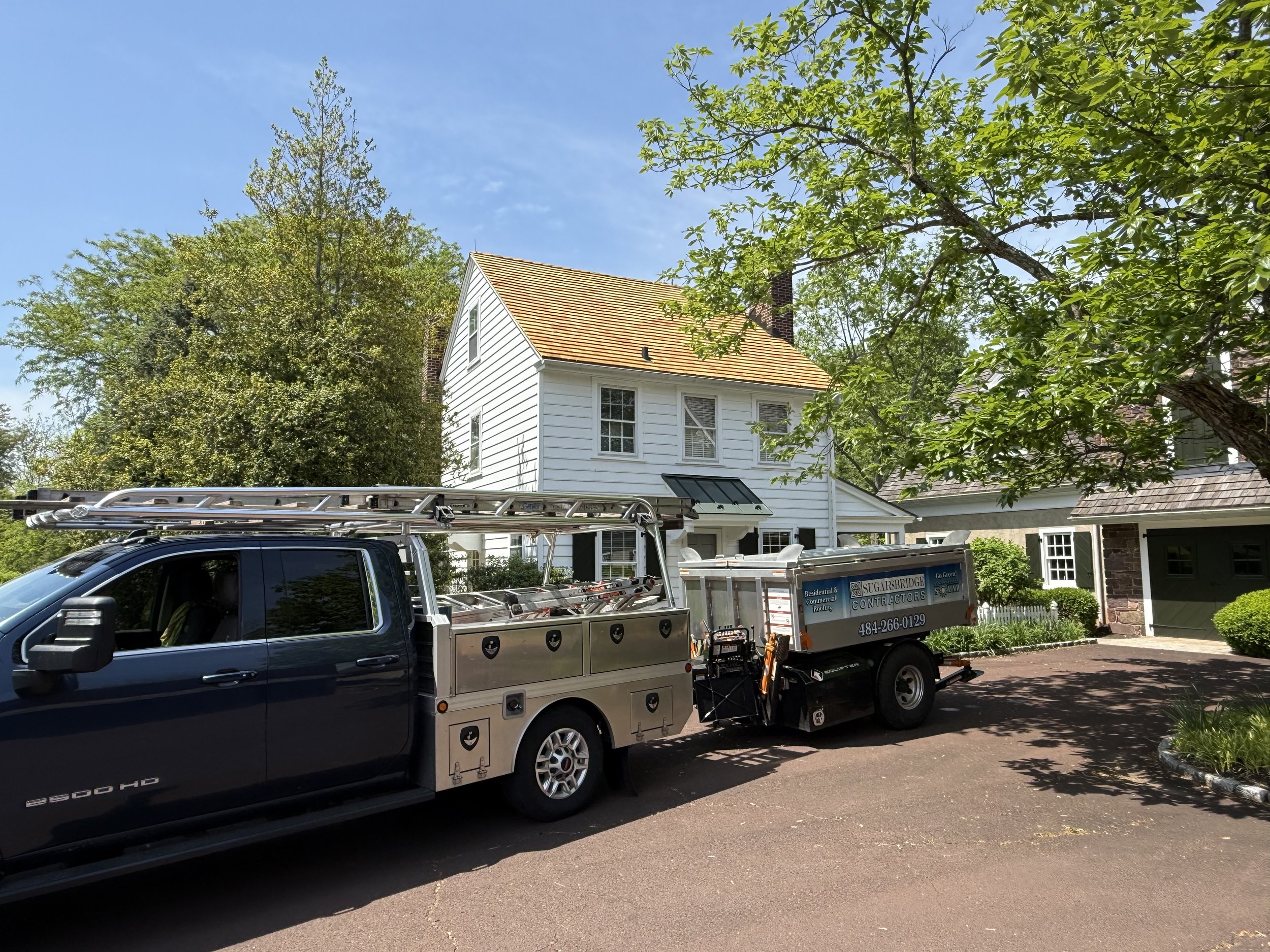 A black utility truck with a ladder on top is parked in a residential driveway, in front of a white house with a sloped yellow roof and some trees.