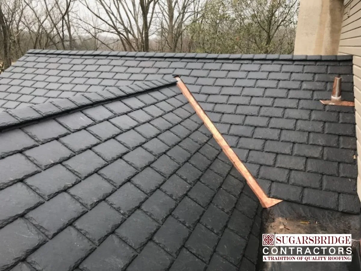 Newly installed black asphalt shingles on a residential roof with wooden rakes, seen from a rooftop view, with trees in the background and a beige house wall on the right.