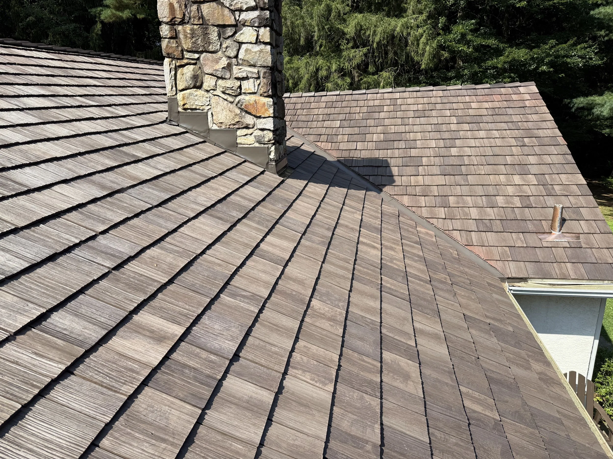 Multiple shingle roofs on a house, including a chimney made of stone and metal flashing, surrounded by trees in the background.