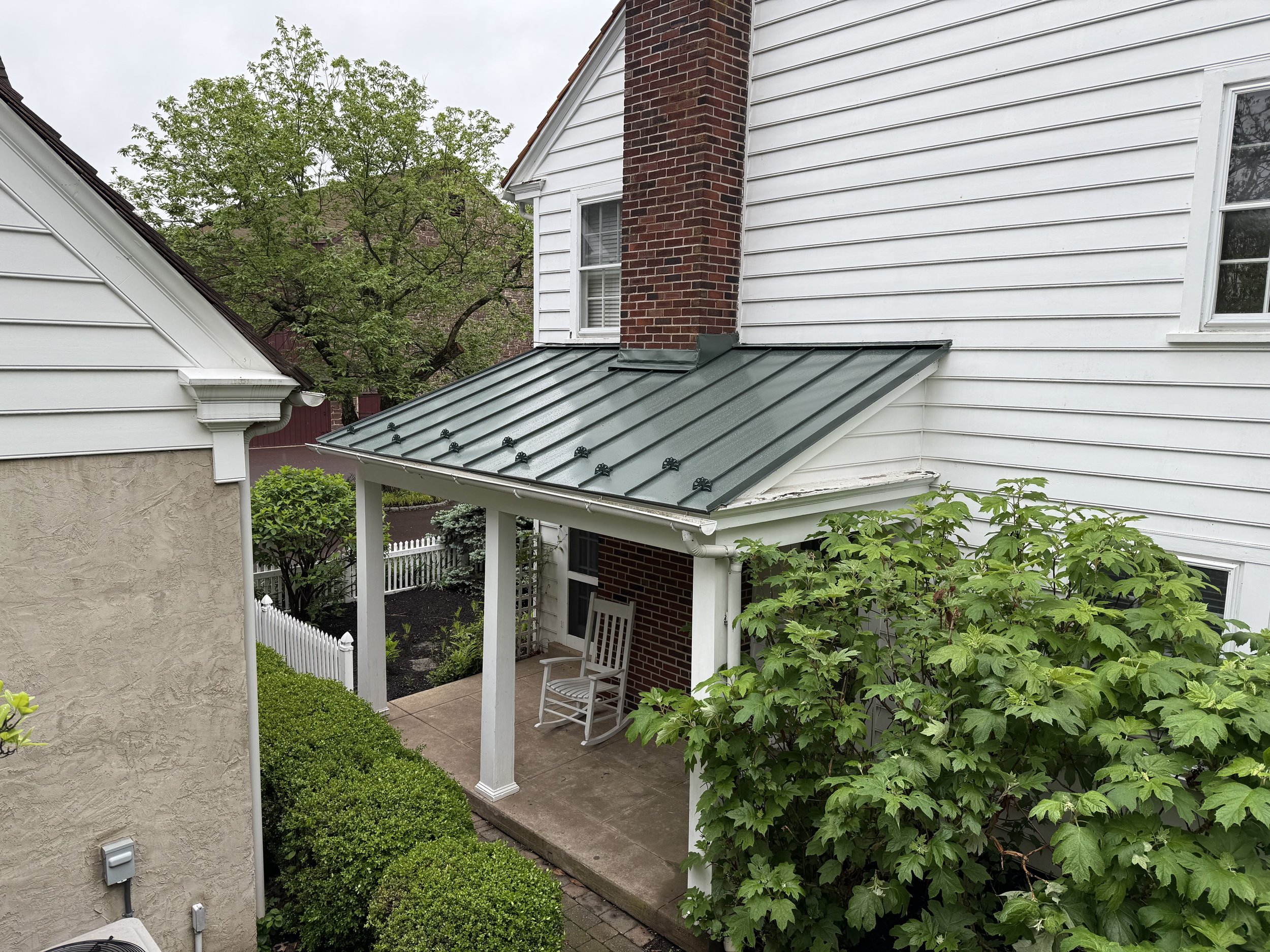 A small covered patio with a white rocking chair, surrounded by lush green bushes and trees, located between two houses in a suburban neighborhood.