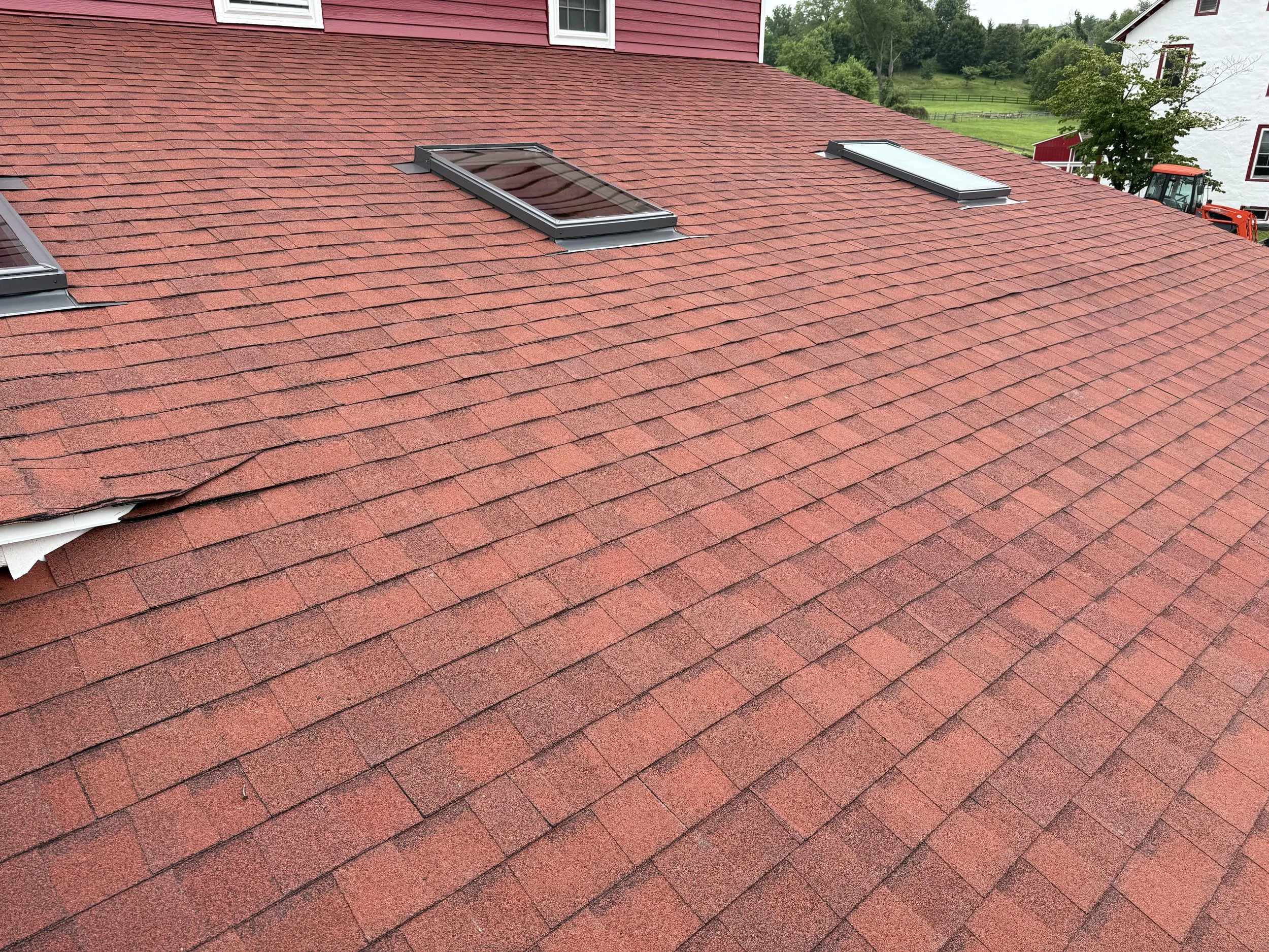 Red asphalt shingle roof with three skylights, one missing some shingles, and a white building with trees and a tractor in the background.