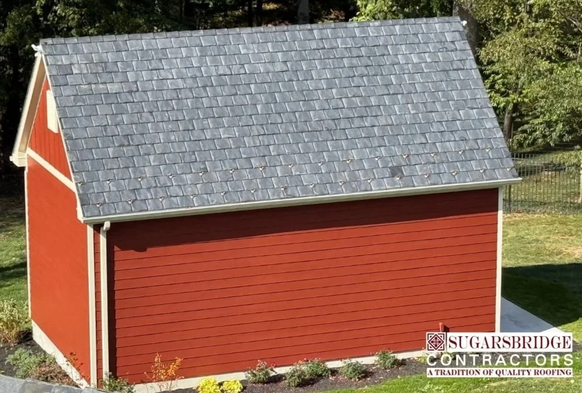 Red shed with a grey Brava Slate Tile roof and small plants at its base, with trees in the background.